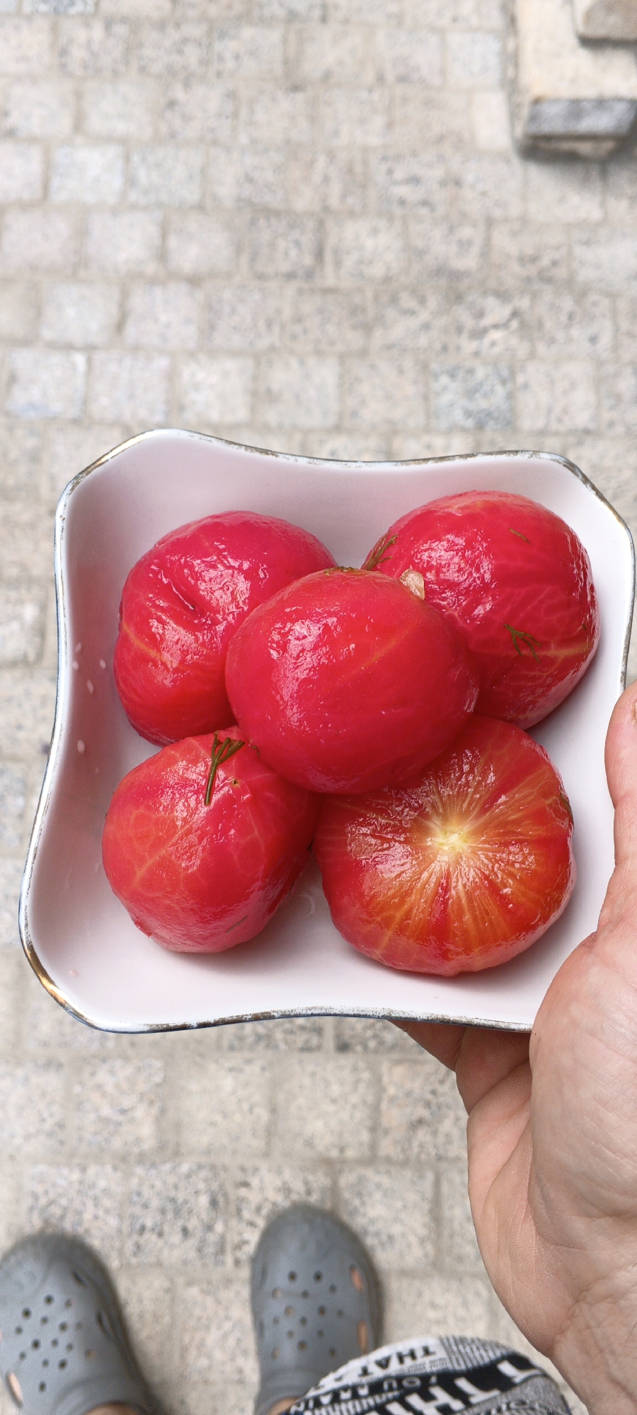 A high-angle, portrait-oriented, close-up shot shows a person's hand holding a white, irregularly shaped bowl filled with five peeled, bright red tomatoes. The tomatoes are plump and glistening, with visible striations from the peeling process. One tomato on the bottom right is cut open, revealing its juicy, segmented interior. The bowl has a thin gold trim and a delicate white dotted pattern on its interior. The background consists of a light-colored, textured stone or brick pathway. The person's feet are visible at the bottom of the frame, wearing grey croc-like shoes and patterned pants. The image does not provide specific clues to identify the location as Tashkent, Uzbekistan.