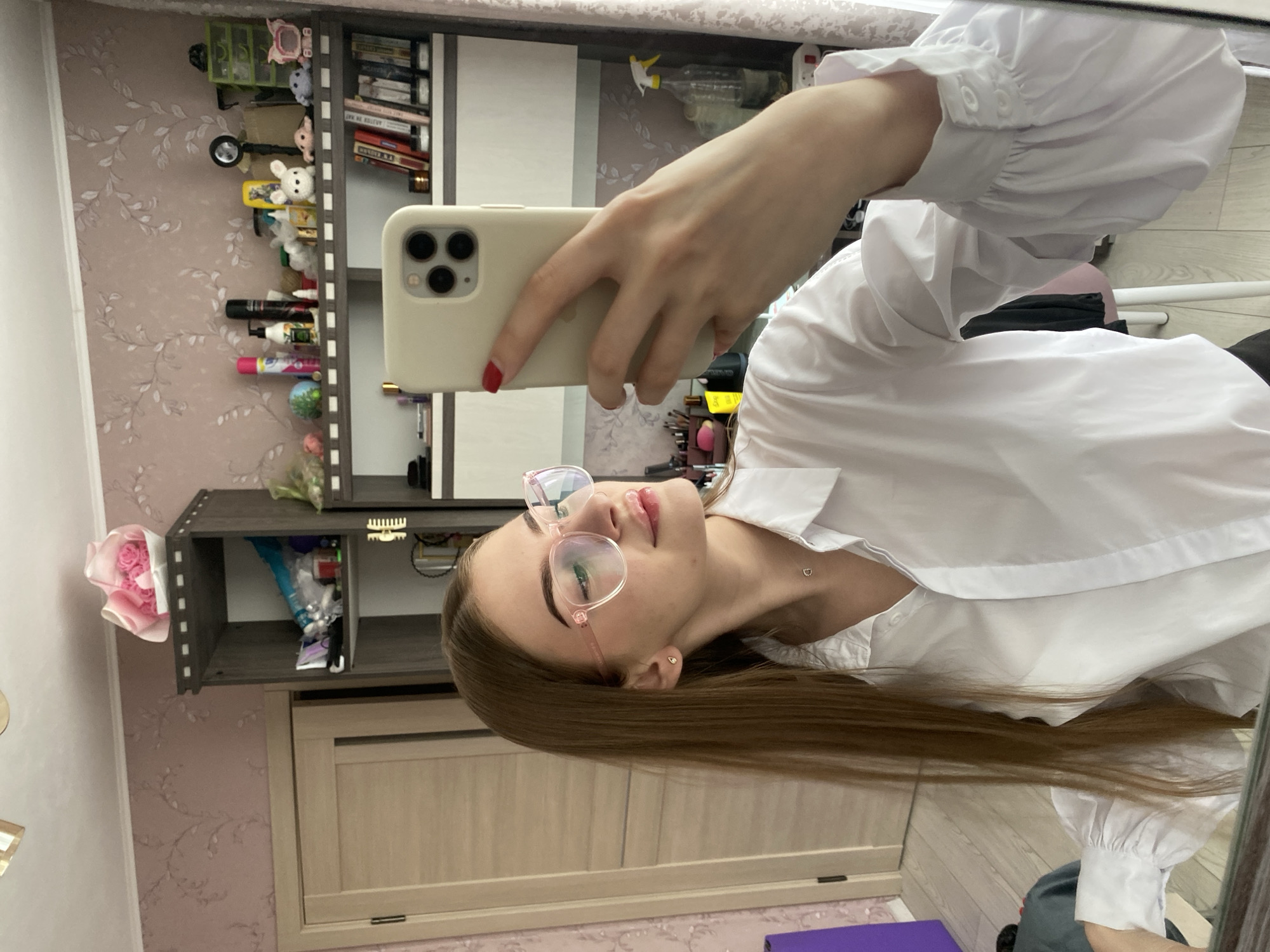 A young woman with long brown hair and pink-rimmed glasses is taking a selfie in a room. She is wearing a white collared shirt with voluminous sleeves and a delicate necklace. The background shows a bookshelf filled with various items, including stuffed animals, toiletries, and books. A door is visible to the left, and a patterned wallpaper adorns the walls. The overall impression is one of a casual, personal moment captured in an indoor setting, possibly a bedroom or living area. There is no indication of the specific location in the city of Dyakovo, Russia.