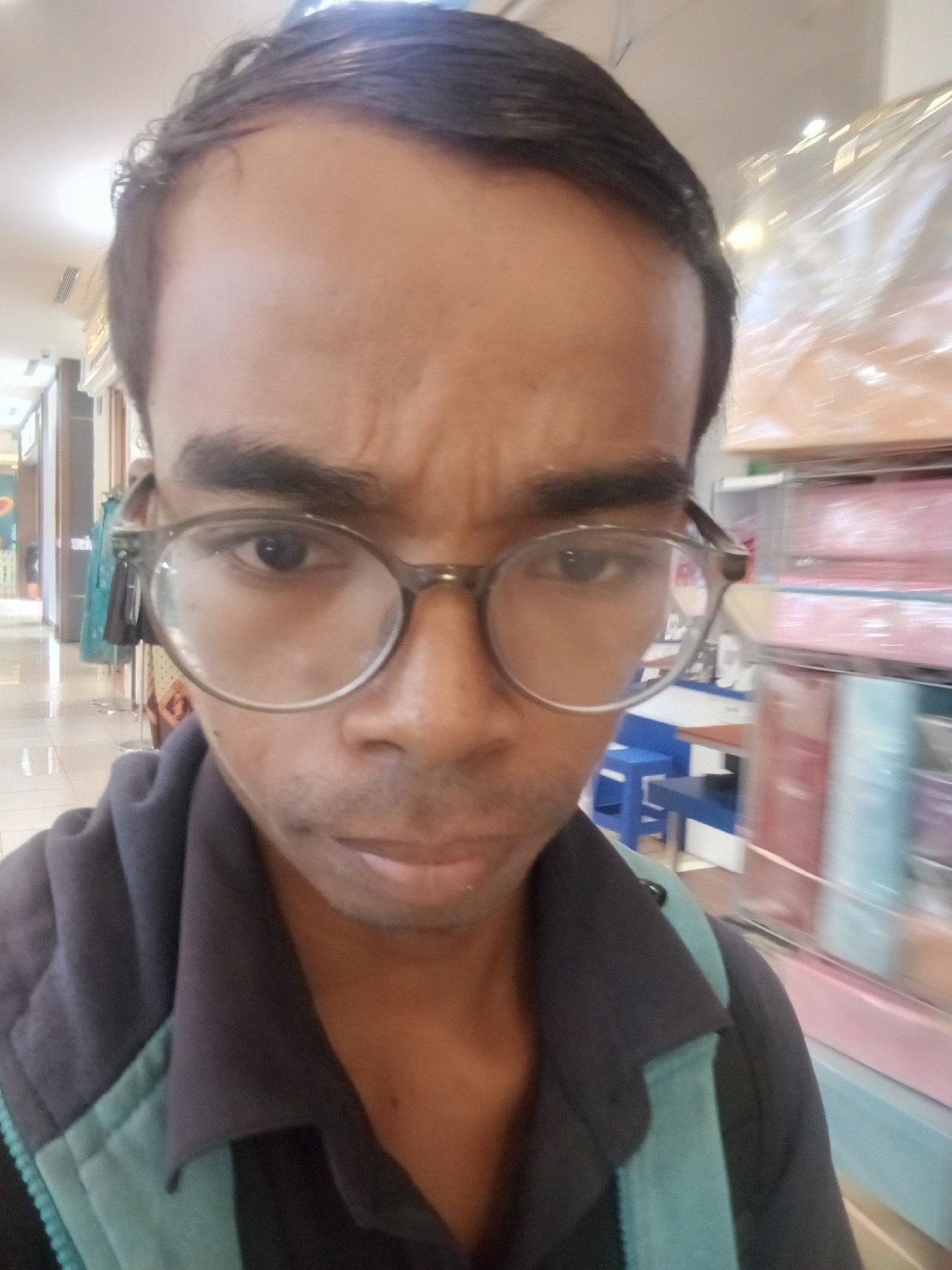 The image is a close-up selfie of a man wearing glasses and a dark jacket with teal accents. He has dark hair and his eyebrows are furrowed, giving him a concerned or thoughtful expression. The background appears to be an indoor setting, possibly a mall or a store, with shelves displaying various items, some of which are wrapped in plastic. The lighting suggests an indoor environment. The man is looking directly at the camera, and his face fills most of the frame. There is no discernible information in the image to specifically link it to Surabaya, Indonesia.