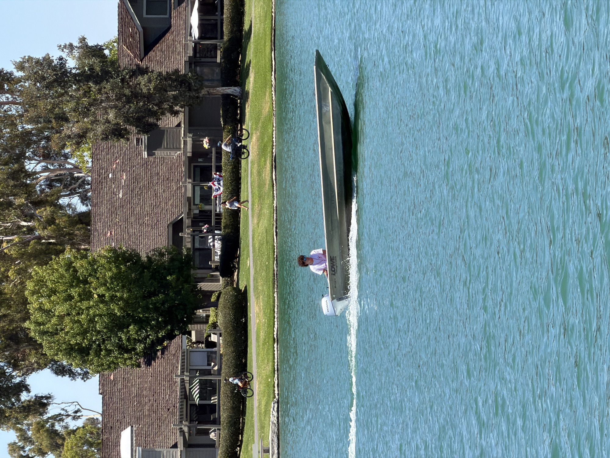 A young person with curly hair and sunglasses is operating a flat-bottomed boat with a Honda outboard motor across a body of water in Irvine, California. The boat is leaving a white wake. In the background, a building with a shingled roof and multiple windows is visible, bordered by a lawn and trees. Two people are on bicycles in the background, and another person is walking. The sky is clear and blue.