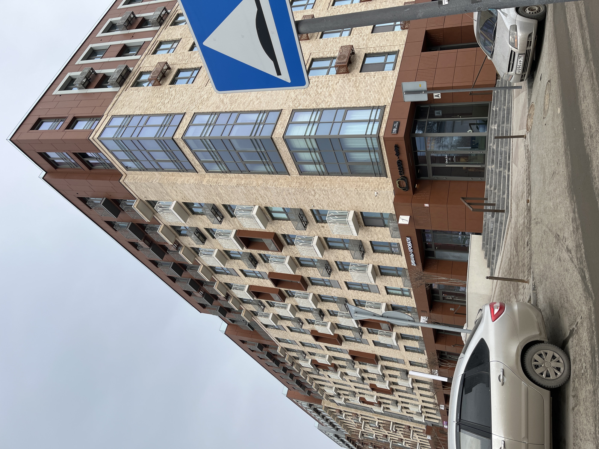The image depicts a modern, multi-story residential and commercial building located at Ulitsa Maksima Gor'kogo, 210, Izhevsk, Udmurt Republic, Russia, under an overcast sky. The building's facade is constructed with light-colored brick on the lower and middle sections, transitioning to reddish-brown paneling on the upper floors and ground-level commercial frontages. Numerous balconies with white or grey metal railings are visible across the building's face, with some units featuring prominent corner bay windows. The ground floor houses commercial establishments, identifiable by signage including "CLOUD-design" and "Диваны-Кровати" (Sofas-Beds), accessed via outdoor steps.
In the foreground, two silver sedans are parked on the paved street or parking area. A silver vehicle, possibly a Lada Granta, is positioned on the left, showing its rear-left side. A second silver sedan, likely a Chevrolet Aveo, is partially visible on the far right, displaying Russian license plate A694YP 18. A blue rectangular road sign, indicating a speed bump with an inverted 'U' graphic, is prominently displayed on a metal pole in the right foreground. The paved ground surfaces show traces of dirt and minimal patches of slush or light snow. No individuals are present, and no direct actions or interactions are depicted within the frame.