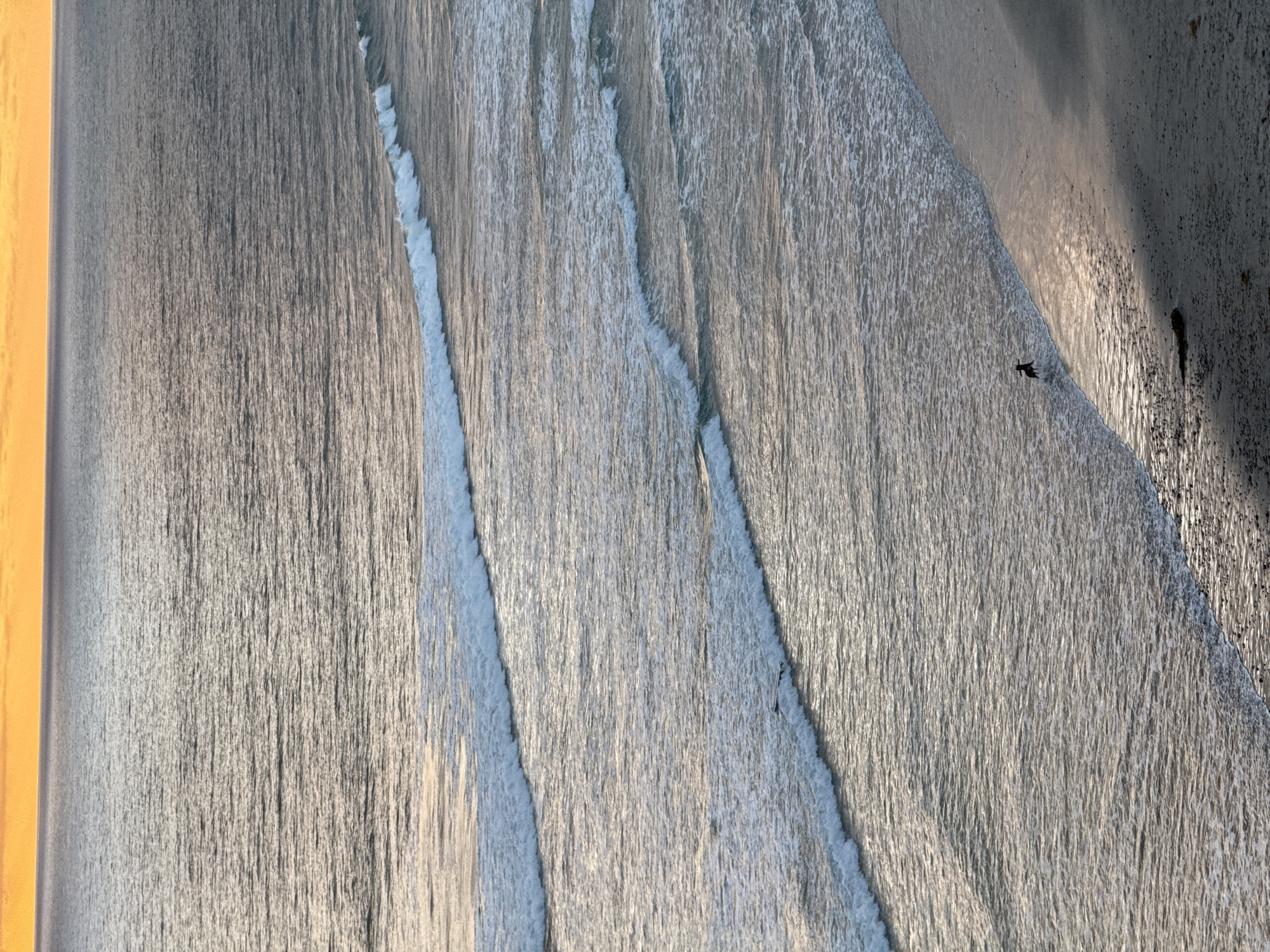 An aerial view captures a section of coastline at dusk in Encinitas, California, USA. The horizon is illuminated by a gradient of orange and yellow hues, indicating the setting sun. The ocean, rendered in muted grays and blues, exhibits distinct parallel wave patterns. Three prominent wave lines with white crests are visible, moving towards the sandy shore. On the beach, a lone figure, appearing as a silhouette, stands at the edge of the water, with one leg submerged. The beach itself is a mix of sand and small pebbles, with areas of shadow and sunlight.