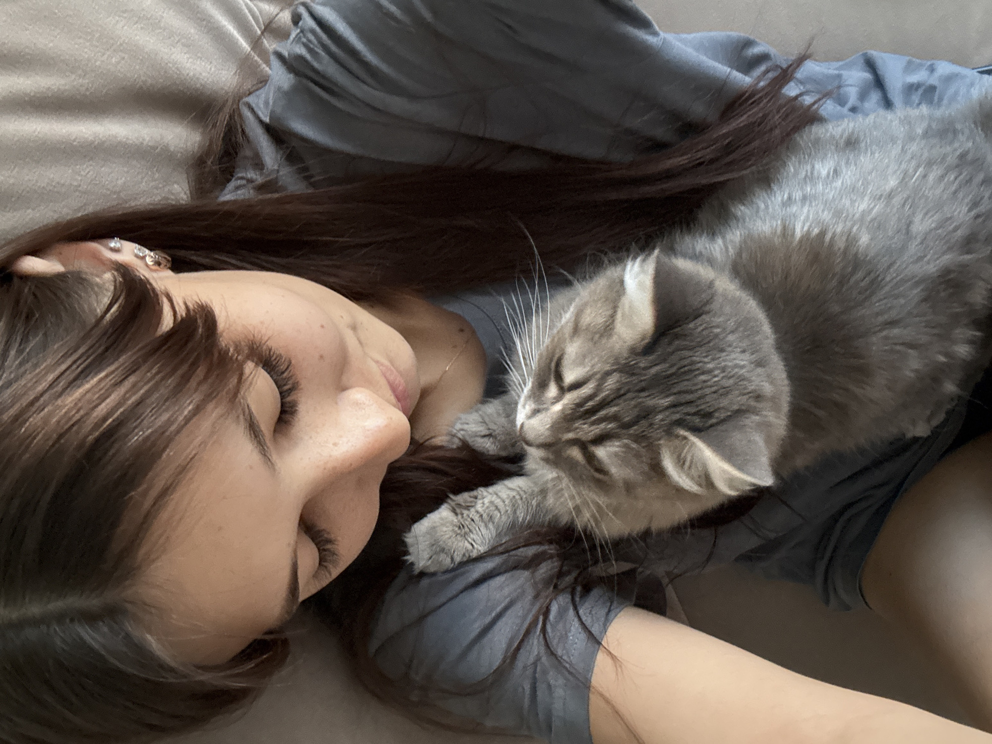 A young woman with long, dark brown hair and long eyelashes is lying down, holding a grey tabby cat. The woman is wearing a grey t-shirt. The cat is nestled in her arms, facing upwards towards her face. The background is a blurred brown surface, possibly a couch or bed. There is no visible indication of the location being Novokuznetsk, Russia.