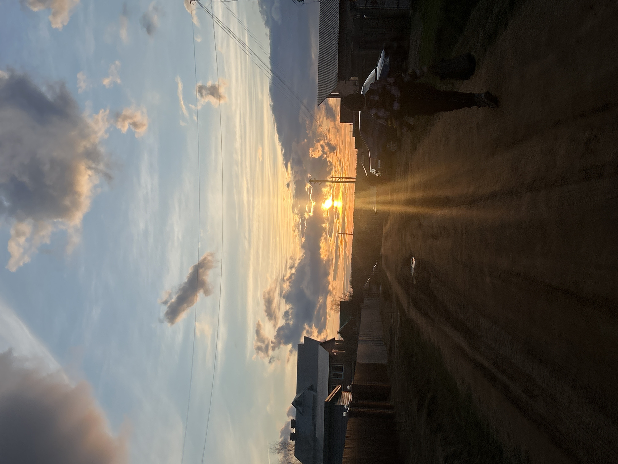 The image captures a serene rural or semi-rural scene in Olkhovka, Russia, at either sunrise or sunset. The sun, a brilliant golden sphere, is positioned low on the horizon towards the right side of the frame, emanating strong, radiant beams that stretch across the sky. The light illuminates a band of clouds, transforming them into striking streaks of orange, yellow, and gold, while other clouds appear darker and more shadowed against the bright backdrop. The sky transitions from a soft blue at the top to warmer hues closer to the horizon.
A dirt road dominates the lower portion of the image, showing visible tracks. On the right side of this road, a person stands mostly in silhouette, appearing to wear a patterned shirt or jacket. Behind the individual, a dark-colored car is parked, also largely silhouetted by the intense light.
Along both edges of the road, several houses or buildings are visible, presented as dark forms against the vibrant sky. Utility poles and overhead wires crisscross the upper left quadrant of the sky, indicating basic infrastructure. The overall impression is one of peaceful natural beauty, with the dramatic light of the rising or setting sun serving as the focal point. No text is visible in the image.