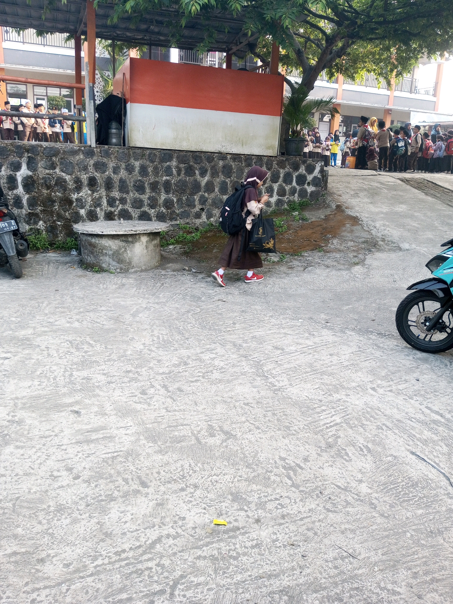 In Krasak, Indonesia, a young student in a brown uniform and hijab, carrying a backpack and a black tote bag, walks across a concrete courtyard in front of a building with a red and white facade. A stone wall borders the courtyard, and a round concrete well is visible near the student. Other students are gathered in the background near the building. A motorcycle is parked on the right side of the frame.