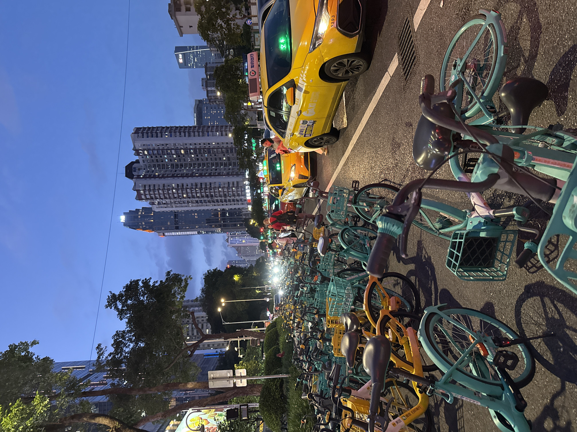 The image shows a street scene in the Tianhe District of Guangzhou, China, at dusk or early evening. The foreground is filled with numerous parked shared bicycles, predominantly in teal and yellow. A yellow taxi is partially visible, situated on the right side of the image along the road. In the middle ground, more taxis and pedestrians can be seen. High-rise buildings form the backdrop, with some illuminated windows indicating residential or office activity. Streetlights illuminate the road and surrounding area. The sky is a gradient of blues, suggesting the transition from day to night.