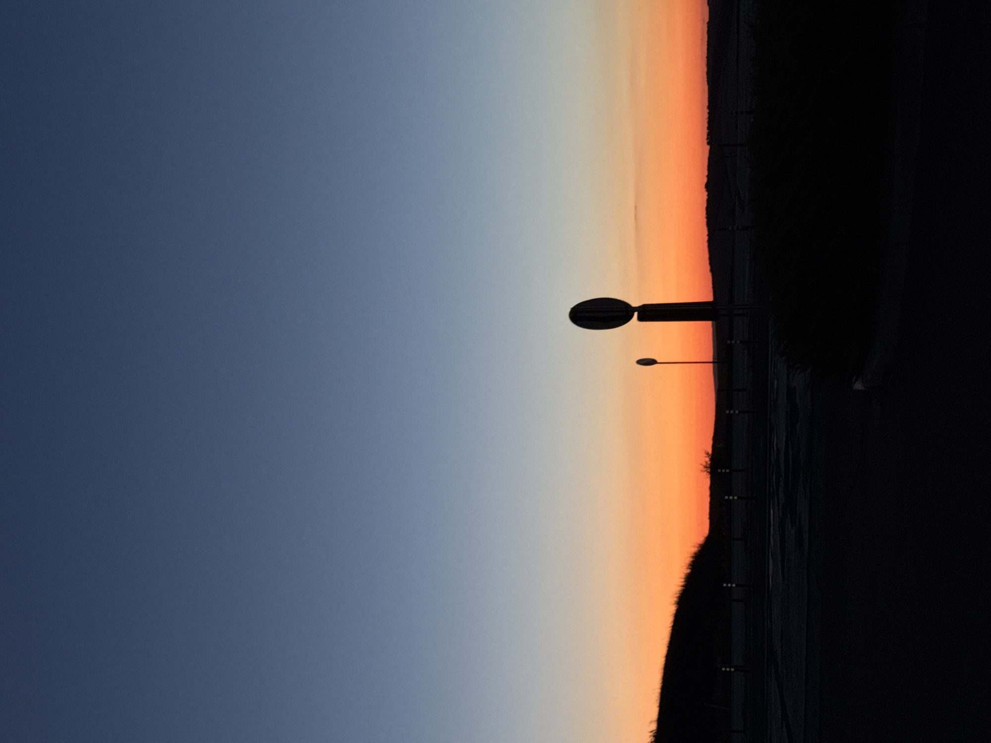 The image depicts a silhouetted landscape at dusk with a clear gradient sky, transitioning from deep blue at the top to vibrant orange near the horizon. A dark, undulating hill is visible in the background, sparsely dotted with small, indistinct trees. In the foreground, a dark, paved area, possibly a road or parking lot, is visible, with a line of small, reflective posts running across it. Two tall, dark poles with circular, silhouetted signs stand on the right side of the frame, one significantly larger and closer than the other. The overall scene suggests an open, possibly rural or semi-rural area outside of the city of Porumbrei, Moldova, given the context. No people or discernible activities are present.