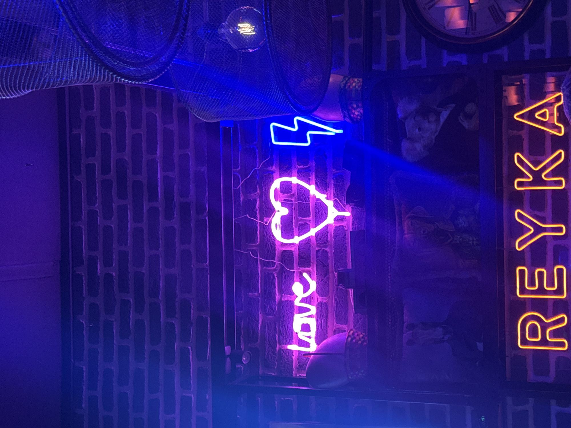 This media file captures a vibrant, neon-lit interior scene, likely a bar or lounge, characterized by a dark, textured brick wall illuminated predominantly by blue and purple light. The setting creates a nocturnal and atmospheric ambiance.
Dominating the mid-ground are several glowing neon signs. A prominent purple-pink neon sign spells out the word "love" in a cursive script, positioned below a smaller, bright blue neon sign shaped like a lightning bolt. To the right, a vertical neon sign spells "REYKA" in a warm yellow-orange hue, standing out against the cooler tones of the other lights.
The dark brick wall serves as the primary backdrop, its texture subtly highlighted by the colored lights. Above the neon signs, a dark, mesh-like ceiling fixture or decorative element is visible, with a light bulb partially glowing. A classic clock face with Roman numerals is partially visible in the upper right corner, adding a touch of vintage character. Below the "REYKA" sign and beside the "love" sign, indistinct dark framed images or artworks adorn the wall. No people are visible in the scene, emphasizing the focus on the interior decor and lighting. The overall impression is that of a trendy, inviting venue experienced during the evening hours. While the image itself does not provide specific geographical cues, the context provided indicates the location is Plovdiv, Bulgaria.