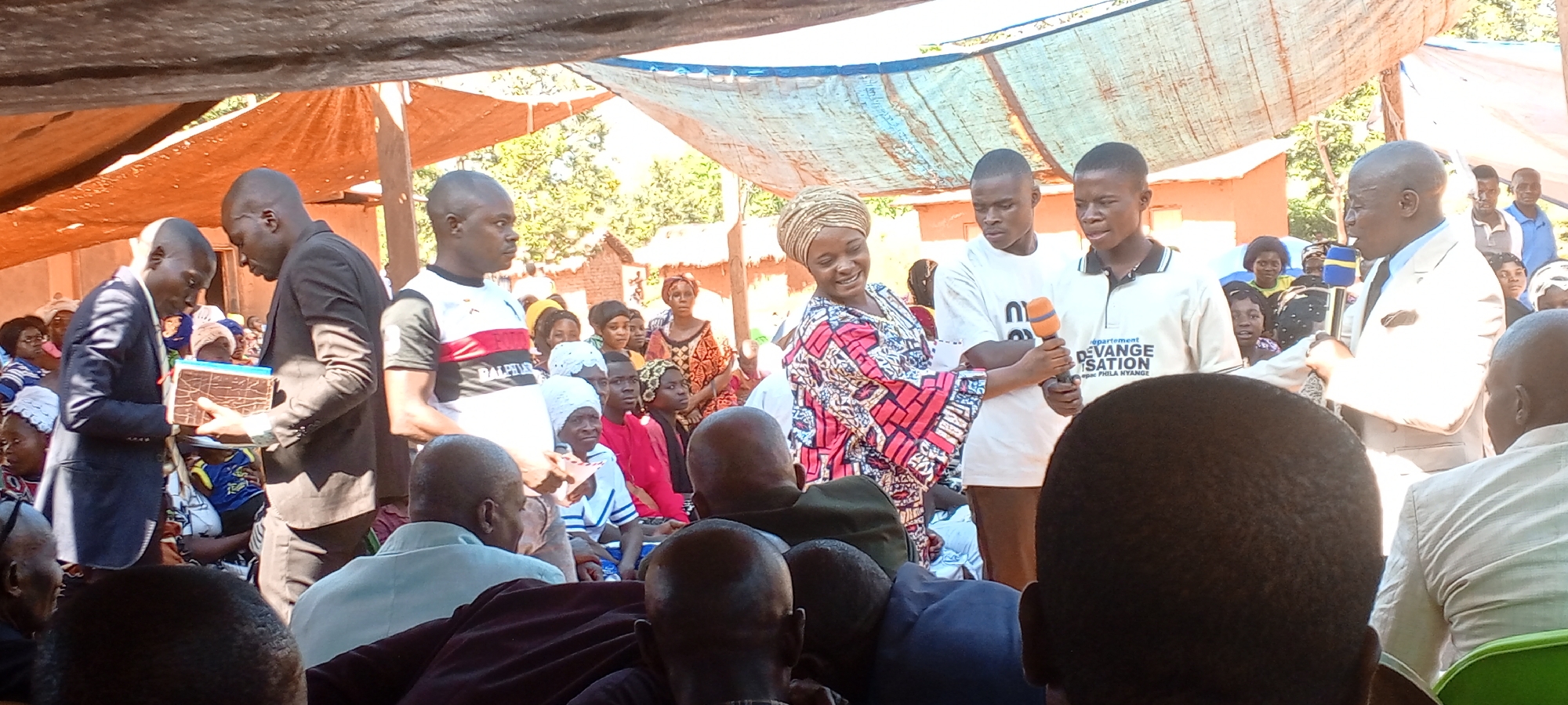 The image depicts a gathering of people in what appears to be an outdoor event in the city of Kalemie, Democratic Republic of the Congo. The crowd is diverse, with men, women, and possibly children present. Some individuals are seated, while others are standing. Several people are dressed in formal attire, including suits and traditional clothing. Some individuals appear to be speaking into a microphone. A tent or makeshift roof provides shade for the event. The environment suggests a rural or semi-rural setting, with trees and rudimentary structures visible in the background.