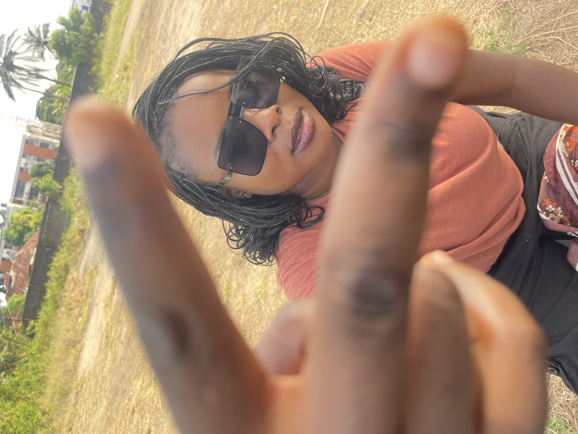 A medium shot captures a Black woman wearing dark sunglasses, a salmon-colored top, and black pants, while making a peace sign with her right hand. The background includes buildings, trees, and grass, suggesting a location in a city or park area.