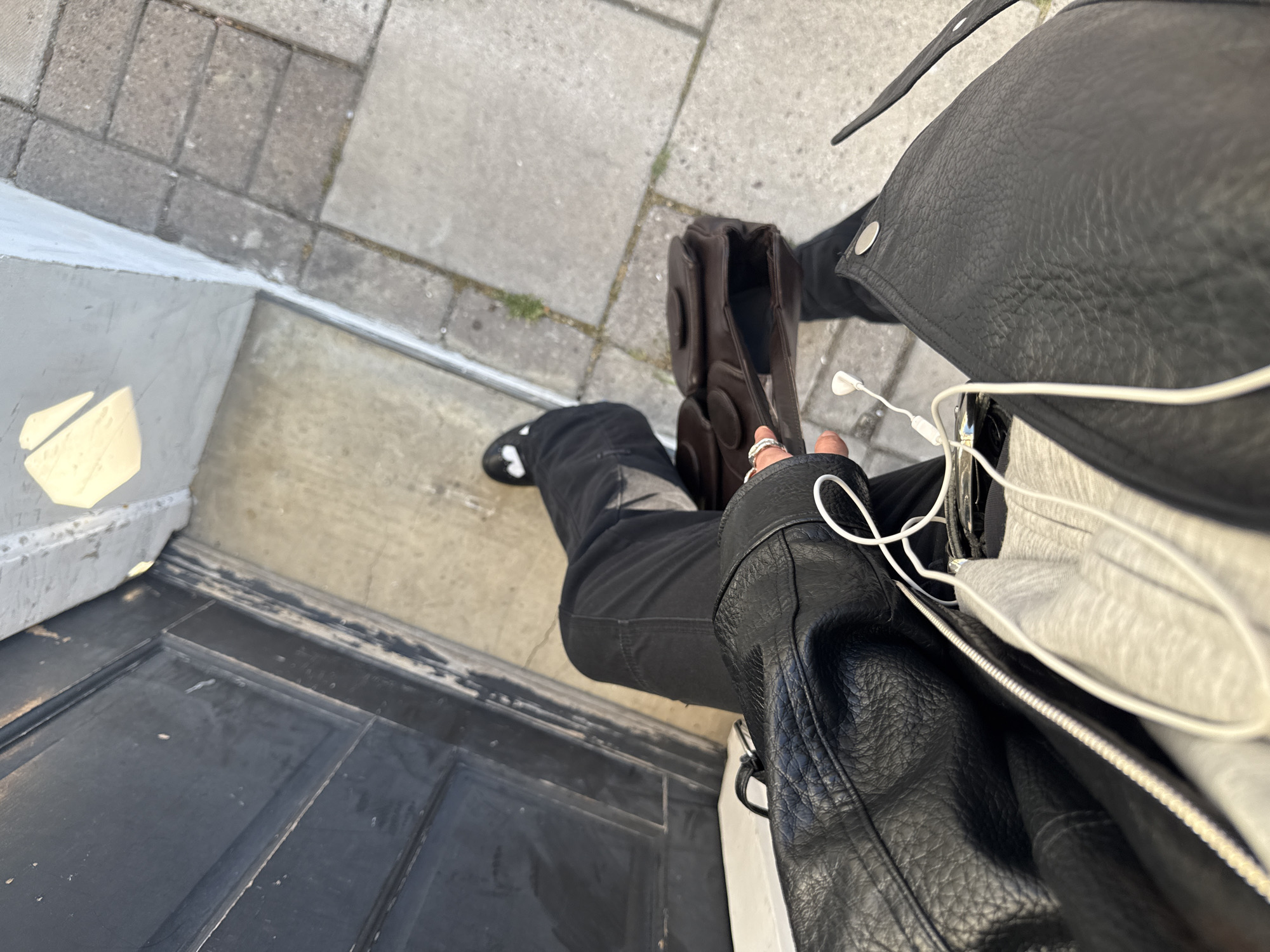 The image captures an overhead, close-up perspective of a person standing or sitting at an outdoor doorstep, likely in London, United Kingdom.
The primary subject is a person, whose torso, arms, and legs are partially visible. They are dressed in dark clothing, including a textured black leather jacket with silver snaps and a zipper, and black trousers. A black shoe with a white detail is visible on one foot. The person's left hand, adorned with rings, is holding a structured brown leather bag with round embellishments on its side. White wired earphones are seen tangled around the jacket, with a white face mask tucked into an opening, possibly a pocket or the bag itself.
The setting appears to be an entranceway. Below the person's feet is a dark, possibly black, wooden door with paneling, which leads out to a light-colored concrete threshold or step. Beyond this, grey concrete paving stones or bricks are visible, indicating an outdoor urban environment. Strong sunlight casts a sharp, angular shadow on the white step, suggesting a bright and clear day, likely morning or afternoon. There is no discernible text visible in the image. The activity appears to be the person either waiting, departing, or arriving, captured in a momentary pause.