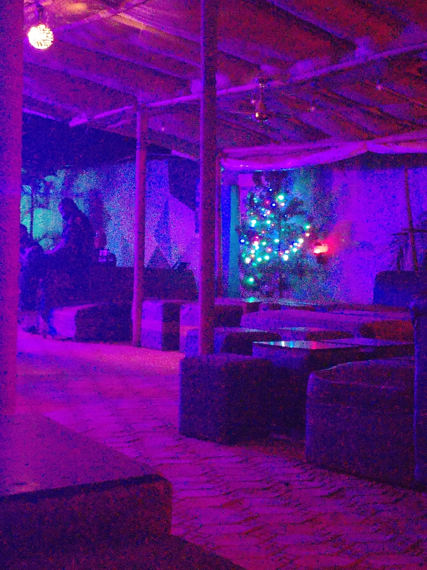 The scene depicts an indoor space decorated with purple and blue lighting, potentially a lounge or bar. Visible structural elements include wooden posts and a roof. Seating is arranged with what appears to be blocky furniture. A Christmas tree with lights is positioned in the background. Two individuals are discernible on the left side of the image. The flooring seems to be made of bricks or tiles. The location is a city of Unknown, Unknown.