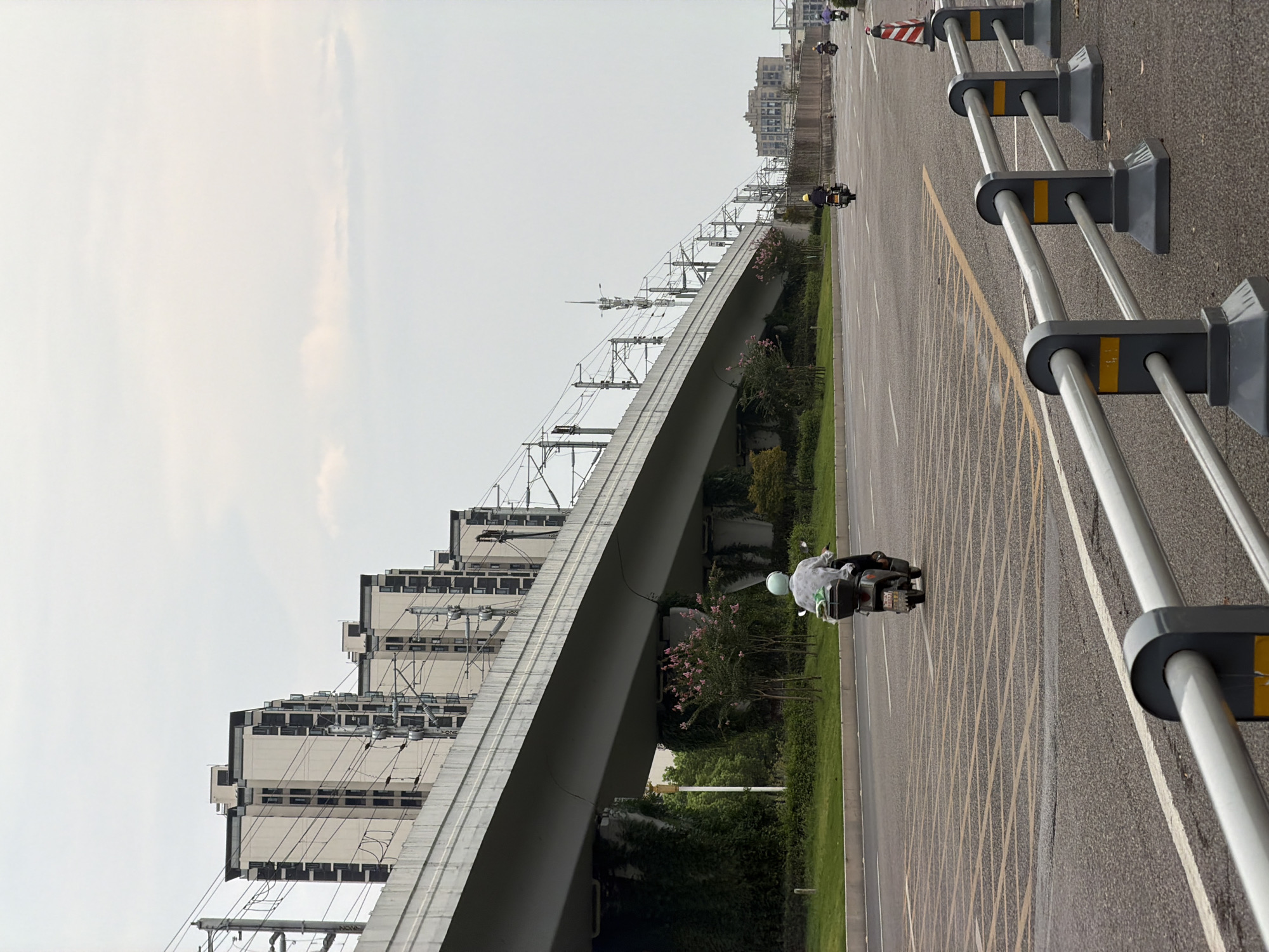 The image depicts a street scene in Jiangjin District, Chongqing, China, on a cloudy day. An elevated train track structure dominates the upper portion of the frame, with several modern residential buildings visible beyond it. The train track is supported by large concrete pillars, some of which are covered in green ivy. Below the train track, a two-lane road is visible, with a person riding a scooter in the foreground, heading away from the viewer. The scooter has a green helmet on the rider's head and a black storage box on the back. Yellow road markings form a grid pattern on the lanes. A metal guardrail runs along the right side of the road, separating it from a sidewalk or embankment. In the distance, other vehicles, including scooters and a car, are also on the road, and more buildings are visible in the far background. A traffic cone is placed on the side of the road in the middle distance. The overall impression is of a developed urban environment with ongoing transportation infrastructure and residential development.
