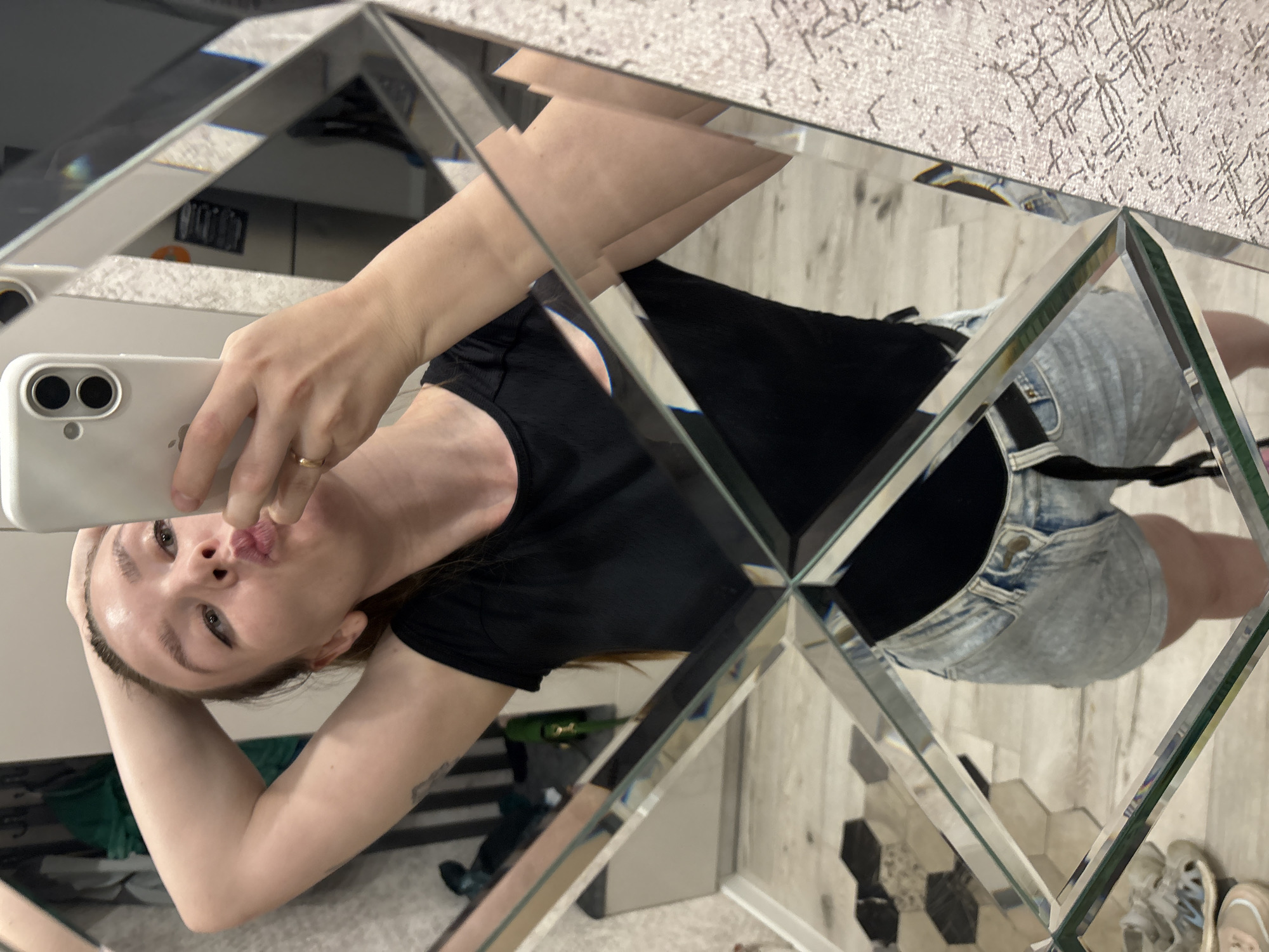 A woman is taking a selfie in a mirror. She is wearing a black t-shirt and denim shorts. Her hair is pulled back, and she is wearing a ring on her finger. She is holding a white smartphone. The mirror is composed of triangular segments. The background is a room with tile flooring, a wall with textured wallpaper, and other furniture. It is unclear from the image whether the location is Izhevsk, Russia.