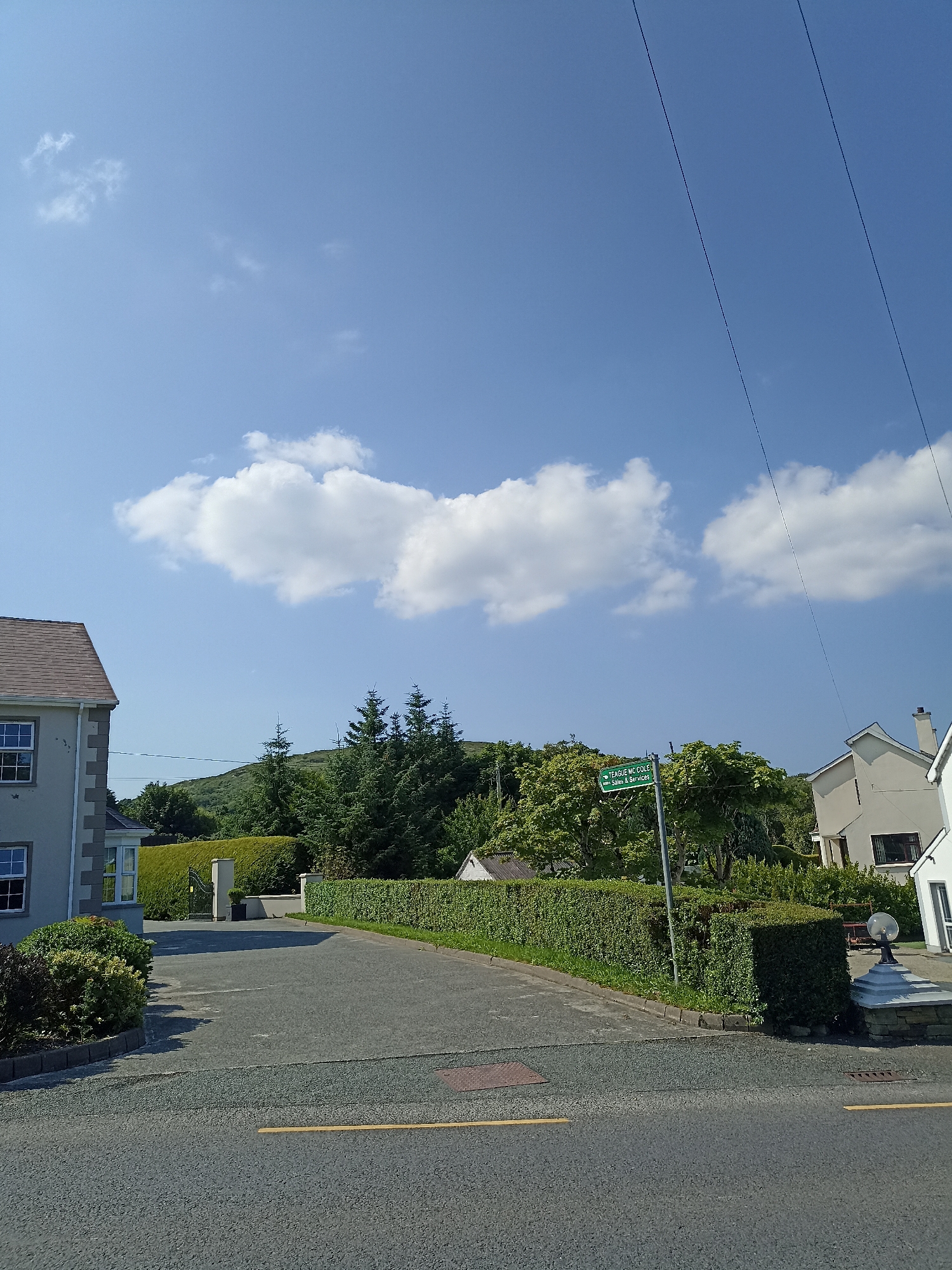 A street-level, eye-level, vertical exterior shot captures a sunny day in Annagry, Ireland, with a bright blue sky dotted with fluffy white clouds. On the left, a grey house with white trim and dark grey roof shingles features multiple windows. A neatly trimmed green hedge forms a boundary around the property. A paved driveway leads away from the house, bordered by more hedges and bushes.
To the right of the driveway, a green street sign is mounted on a pole. The sign reads "TEAGUE MC.COLE" on the top line and "Sales & Services" on the bottom line, indicating a local business or street name. Behind the sign, lush green trees and foliage create a dense backdrop, with a rolling green hill visible in the distance.
A white house with a grey roof and dark windows is partially visible on the right side of the frame, with a decorative stone pillar and lamp post in front of it. Power lines stretch across the upper portion of the image, diagonally traversing the sky. The road in the foreground is paved with asphalt and has yellow lane markings. The scene conveys a peaceful, residential atmosphere on a clear day in the Irish countryside.