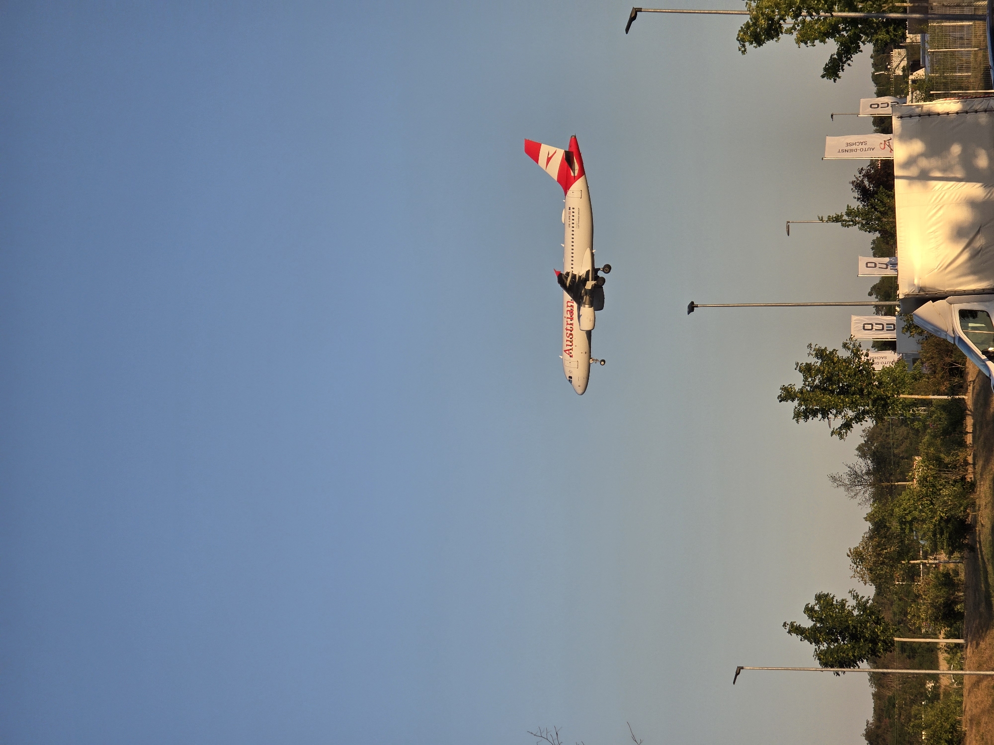An Austrian Airlines Airbus A320 aircraft is captured in mid-flight against a clear blue sky, likely on approach to Frankfurt Airport (FRA) in Kelsterbach, Germany. The aircraft, with its distinctive red and white livery and "Austrian" branding, is positioned in the upper right quadrant of the frame. Below the aircraft, a line of trees and vegetation forms a verdant landscape. Several streetlights are visible in the foreground and mid-ground, interspersed with white banners advertising "ECO" and "AUTO-DIENST SACHSE." A white truck, partially covered with a tarp, is parked in the lower right corner of the image. The overall scene suggests a sunny day with a slight haze, common during the late afternoon or early evening.