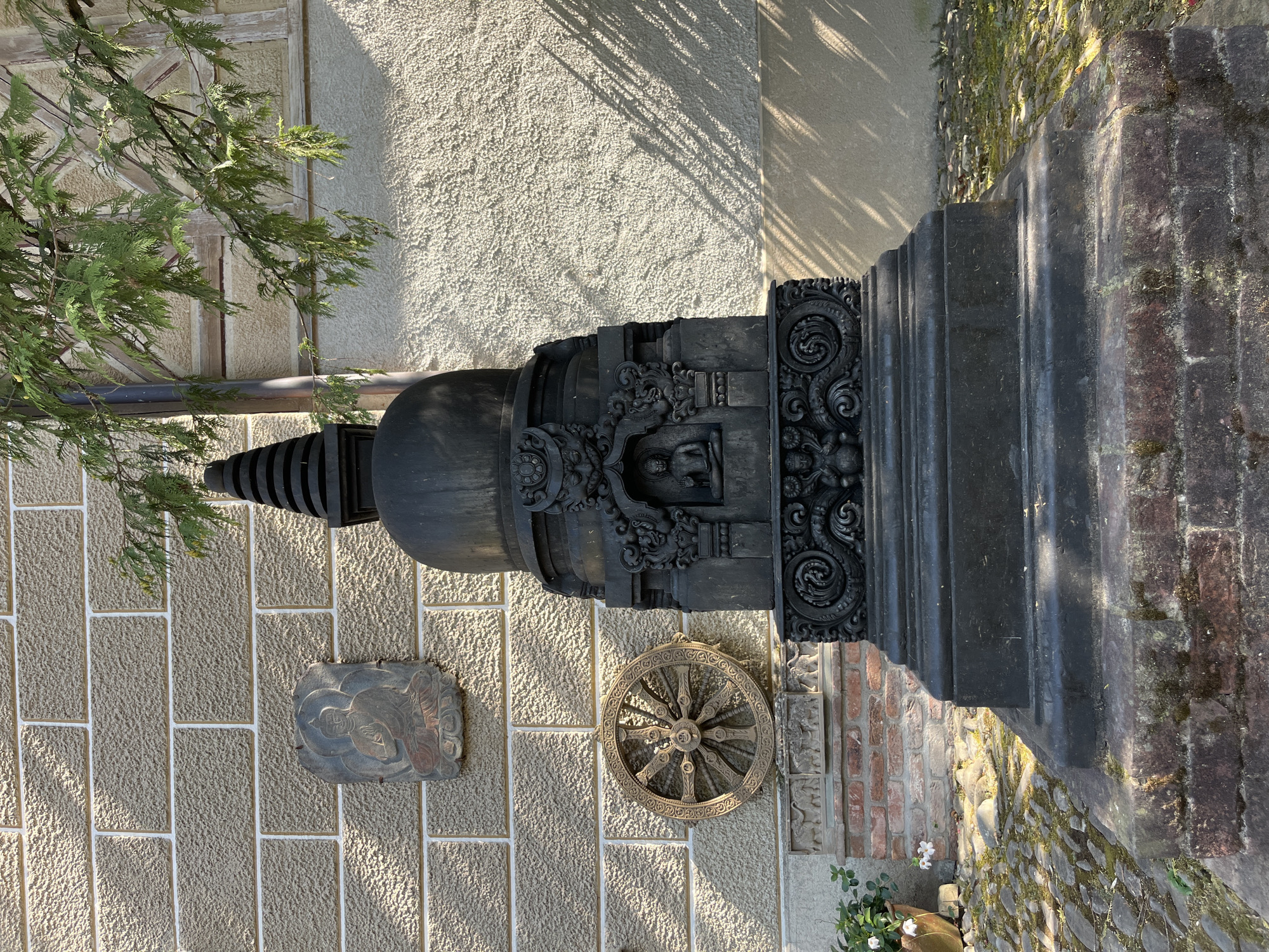 In the city of Epagny, Switzerland, a dark, ornate stupa stands as the central subject of the image, positioned on a base constructed of weathered red bricks. The stupa is elaborately carved with religious iconography, including a central niche containing a depiction of Buddha. To the left of the stupa, mounted on a textured, beige brick wall, are two decorative elements: a bas-relief plaque of a seated Buddha and a wooden wheel with intricate spokes. The wall itself is composed of rectangular, sand-colored bricks with white mortar lines. Below the wall, a cobblestone path, partially covered with moss and fallen leaves, leads towards the stupa. A terracotta pot containing white flowers is visible on the left side of the path. Overhead, branches of a conifer tree extend into the frame, casting dappled shadows on the wall and the stupa. The lighting suggests it is daytime, with sunlight creating strong contrasts.
