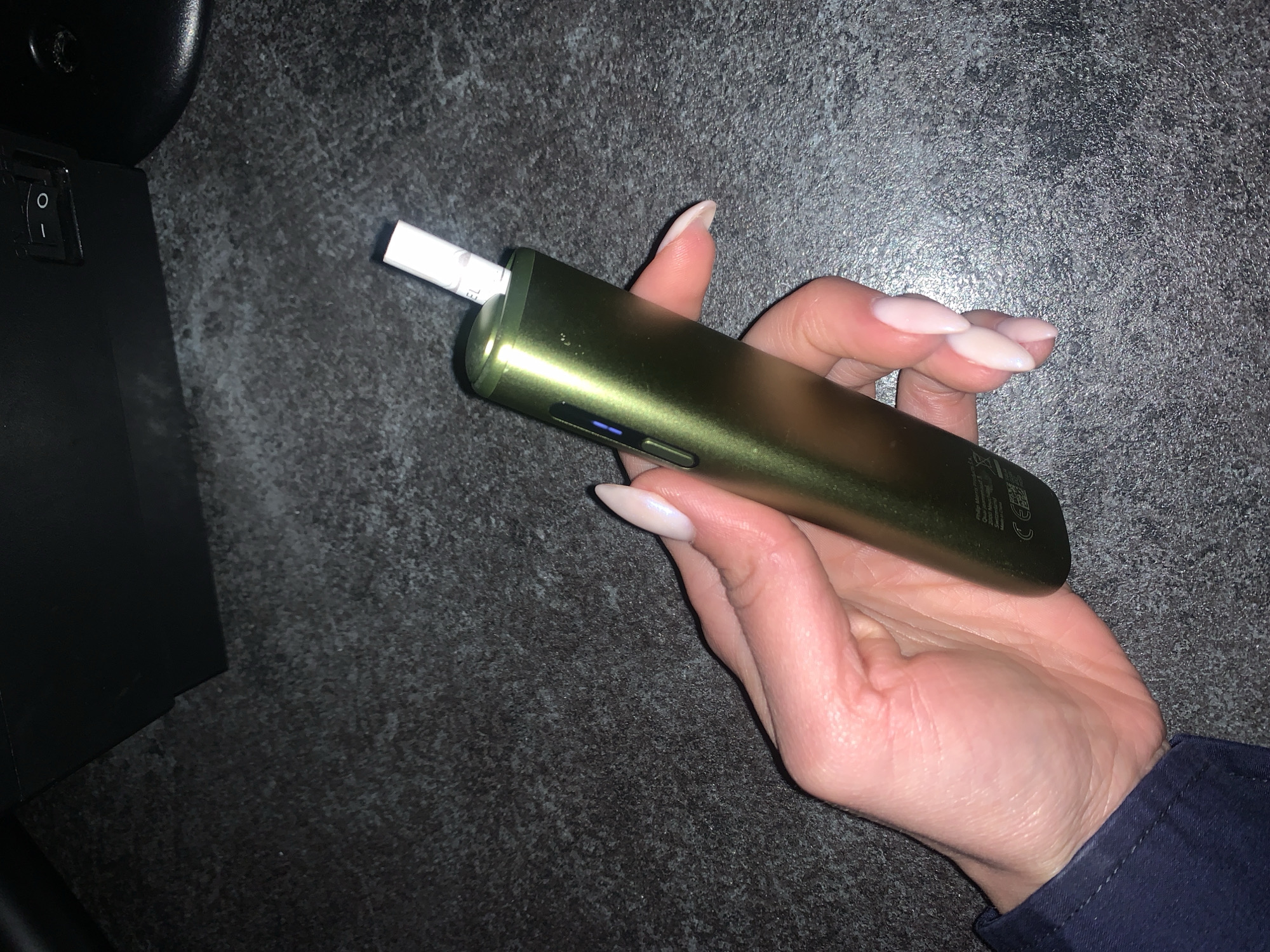 A hand with light-colored skin and pale pink, almond-shaped fingernails holds a metallic green electronic smoking device. A white cigarette-like stick is inserted into the top of the device. The device has a small blue light on its side. The background is a dark, textured surface, possibly a desk or countertop. In the upper right corner of the image, a portion of a black electronic device with an on/off switch is visible. The scene appears to be indoors, in an urban environment, though no specific location within Burgas, Bulgaria, can be identified.