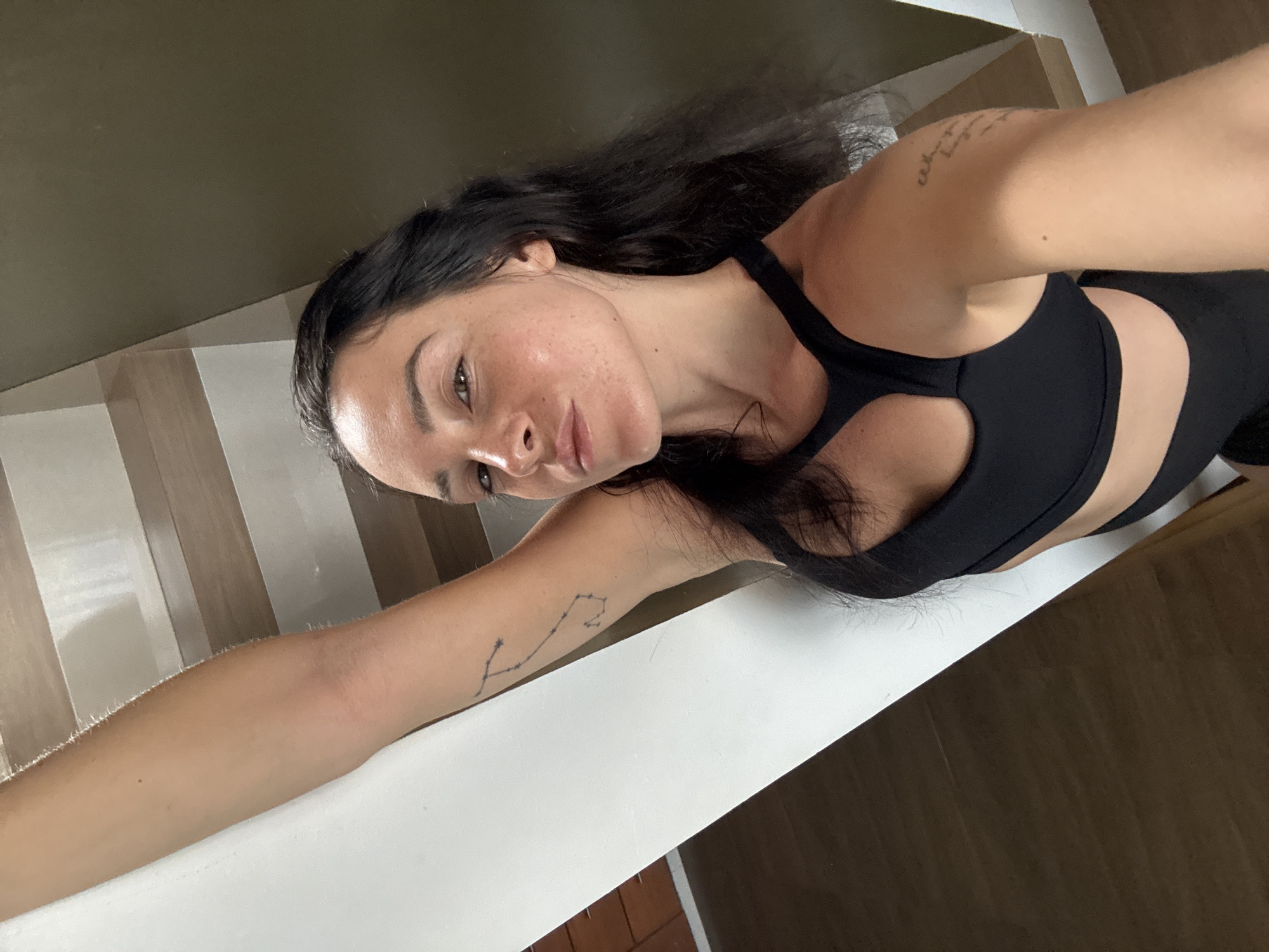 A dark-haired woman with freckles, wearing a black sports bra, poses for a selfie. Her right arm is extended upward, resting on a white railing. She has a constellation tattoo on her left arm and writing on her right arm. The background shows wooden stairs and a wall. The image appears to have been taken indoors, possibly in a residential building in North Kuta, Indonesia.