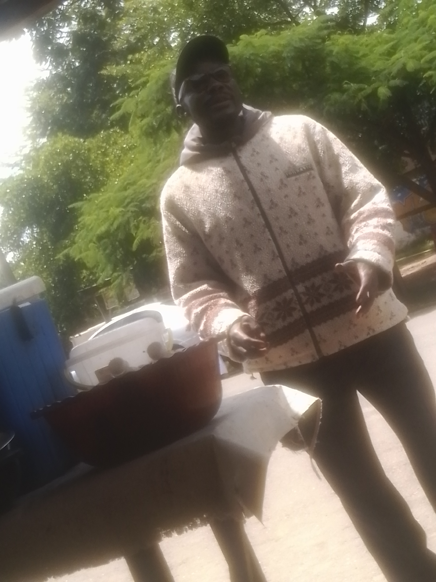 A man wearing a patterned zip-up jacket and a cap stands outdoors in front of lush green foliage. To his left is a blue cooler and a white container on a surface that appears to be a table. In the foreground, a dark-colored bowl is situated on the table. The man is looking towards the left and has his hands near his chest, one hand slightly open. The scene is illuminated by bright, natural light, suggesting it is daytime. The background consists of dense trees and a hint of a wooden structure.