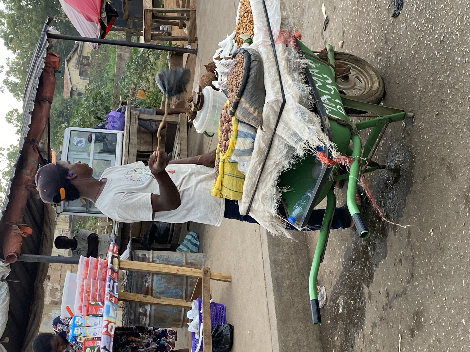 The image depicts a vendor standing beside a green wheelbarrow filled with peanuts and other goods in Bukuru, Nigeria. The vendor, wearing a white shirt, cap, and earpiece, holds a duster. The wheelbarrow is loaded with bags of peanuts and covered with a white cloth. Other market stalls and goods are visible in the background, including a cooler and assorted packaged products. The location appears to be a bustling street or market area.