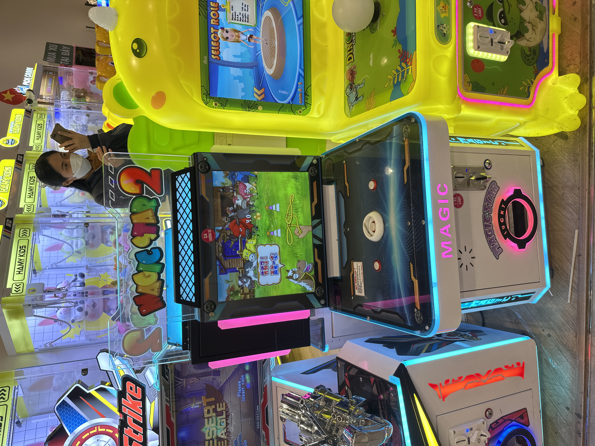 The image depicts a row of arcade games at an indoor entertainment venue, likely a mall or amusement center, in Tay Nha Trang Ward, Vietnam. In the foreground, to the right, is a bright yellow arcade game with a cartoonish dinosaur or frog theme, featuring a screen displaying a character selection interface with the text "SELECT ROLE" and a character named "RIRI". To the left of this yellow machine is a white arcade game with pink and blue neon accents, displaying the title "MAGIC STAR 2" on its screen, which shows a cartoon scene of farm animals and a cowboy. A woman wearing a white face mask is partially visible behind the "MAGIC STAR 2" machine, looking at her phone. To the far left, another arcade game with a "Desert Eagle" theme is visible, along with a chrome-plated gun accessory. The floor is tiled with a wood-like pattern. Other arcade machines with similar bright lighting and cartoon graphics are visible in the background, some appearing to be crane games. The overall environment is brightly lit and appears to be a bustling entertainment area.