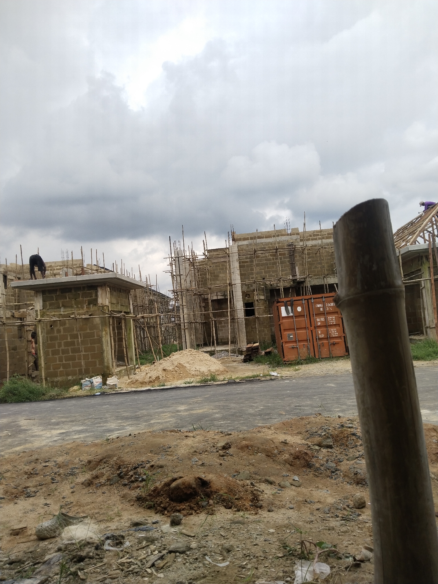 An outdoor, eye-level, medium shot captures a construction site in Uyo, Nigeria, under a cloudy sky. The foreground is dominated by loose soil and debris, with a large section of what appears to be a bamboo pole in the right foreground, partially obscuring the scene. The middle ground features an asphalt road bordering the construction area.
The construction site itself shows several buildings in various stages of completion. The primary structures are made of concrete blocks and are surrounded by scaffolding constructed from bamboo poles. One building in the left middle ground has a person in dark clothing bent over its rooftop, seemingly working. Another smaller building adjacent to it has a figure visible near its entrance, also appearing to be working or observing.
Further into the background, more structures are under construction, also supported by extensive bamboo scaffolding. An orange shipping container is visible on the right side of the middle ground, positioned near one of the buildings. The overall atmosphere is one of active construction, with the natural elements of dirt, grass, and an overcast sky contributing to the scene's raw and unfinished character.