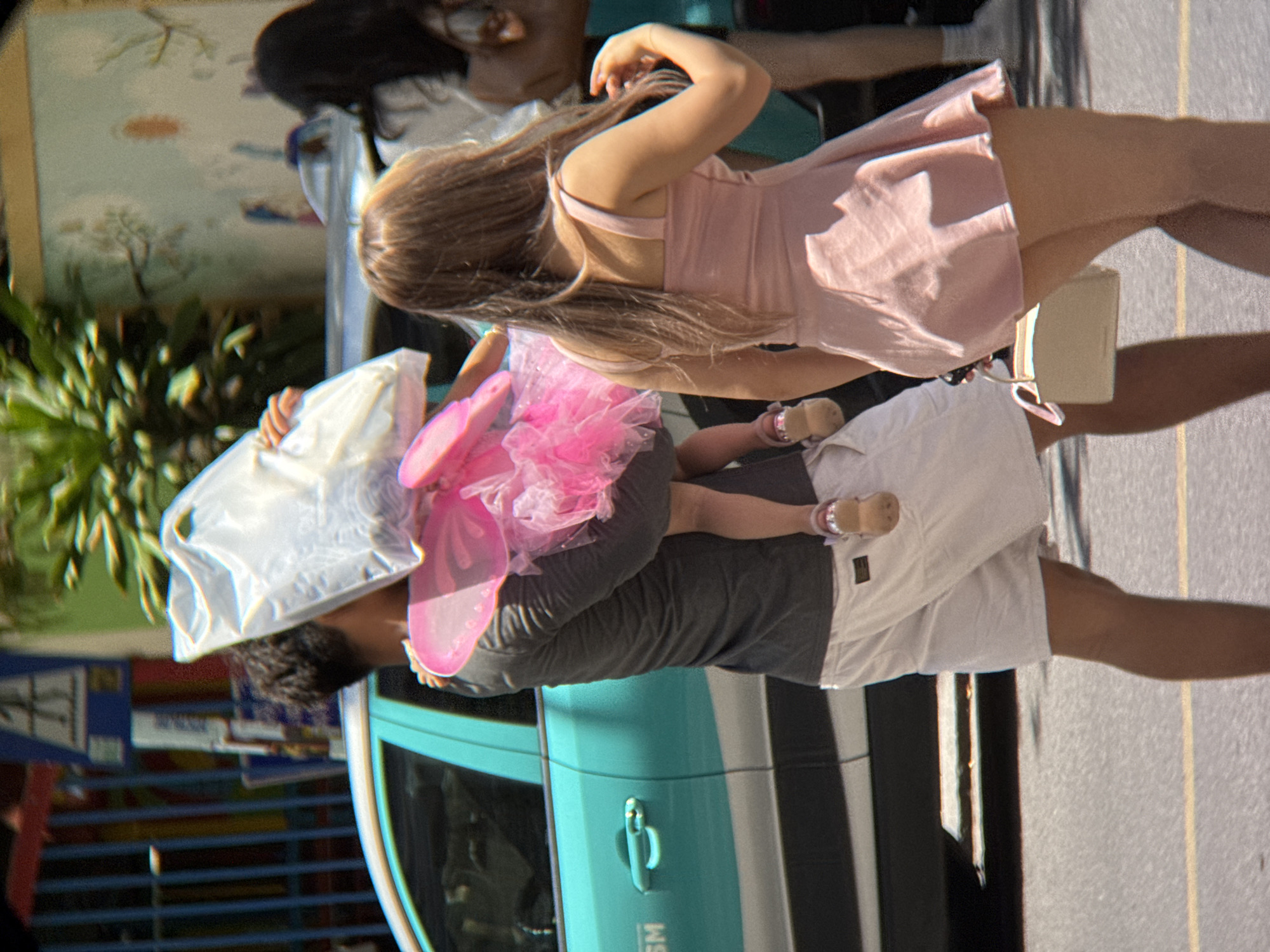 A man, seen from the side, carries a young child dressed in a pink fairy costume. The child is holding a large clear plastic bag. The man is wearing a dark grey long-sleeved shirt and white shorts. He is wearing light brown sandals. A young woman, also seen from the side, walks ahead of him. She has long, light brown hair and is wearing a pink dress and carrying a beige handbag. They are on a paved street. Behind them, a turquoise taxi is parked. The background shows green foliage and a building with a mural. The scene appears to be taking place in Nha Trang, Vietnam, suggested by the style of the taxi and the general environment.