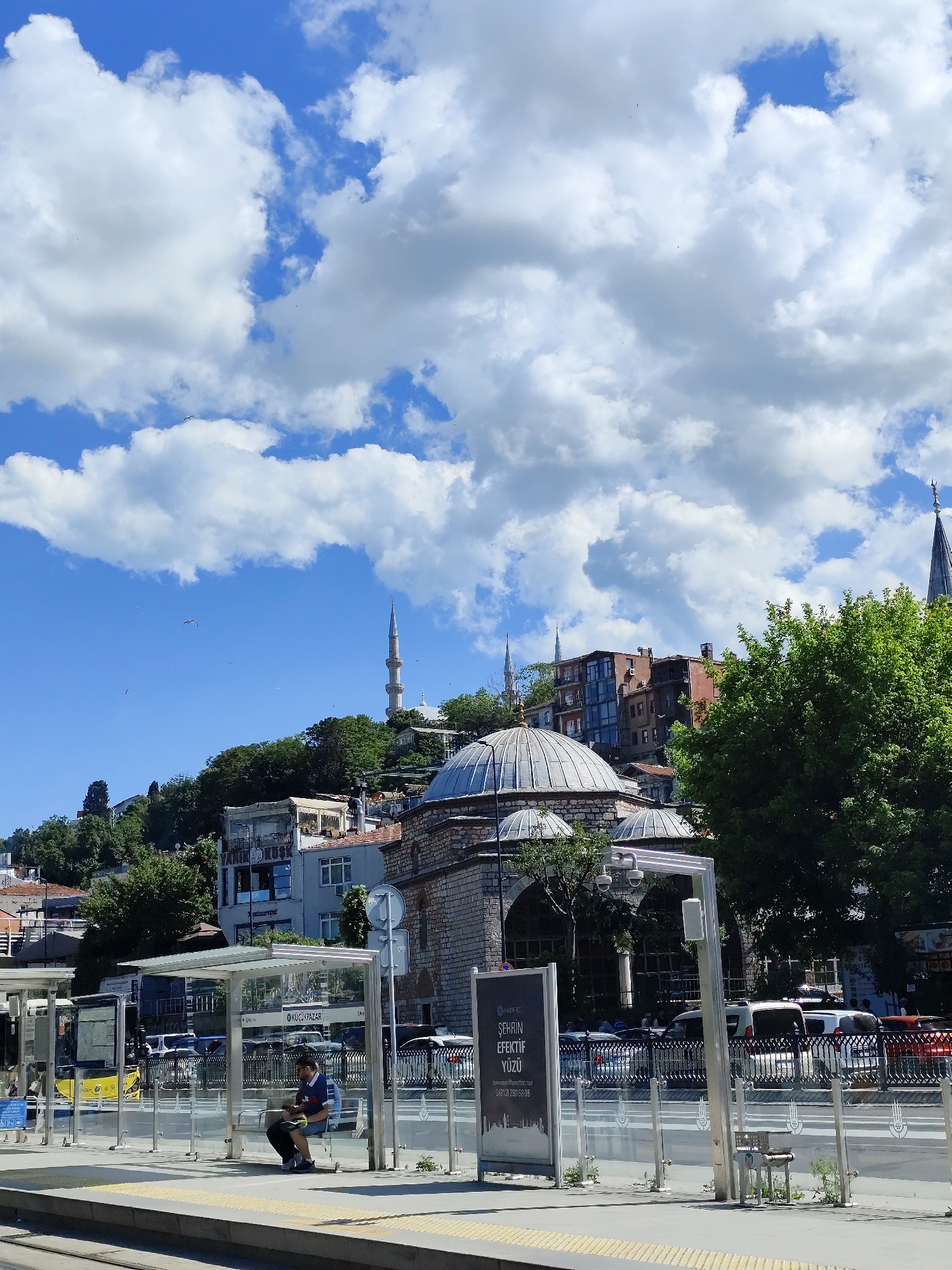 The image captures a street scene on a sunny day in what appears to be Istanbul, Turkey. Fluffy white clouds fill the blue sky, providing a vibrant backdrop to the urban landscape.
In the foreground, there's a modern tram or bus stop shelter made of glass and metal. A man is sitting inside the shelter, seemingly occupied with his phone or another small device.
Behind the shelter, a busy street is visible with cars and other vehicles driving by. An old building with a prominent dome is located behind the bus stop, suggesting a historic structure like a mosque or mausoleum. Further up the hill, there are residential buildings and what appears to be a minaret, reinforcing the impression of Istanbul. A tall spire is on the right side of the frame, indicating the presence of another religious building of a different architectural style.
The atmosphere seems to be lively, typical of a city during the daytime. The image is well-lit and shows clear details, capturing the contrast between old and new elements of the city.