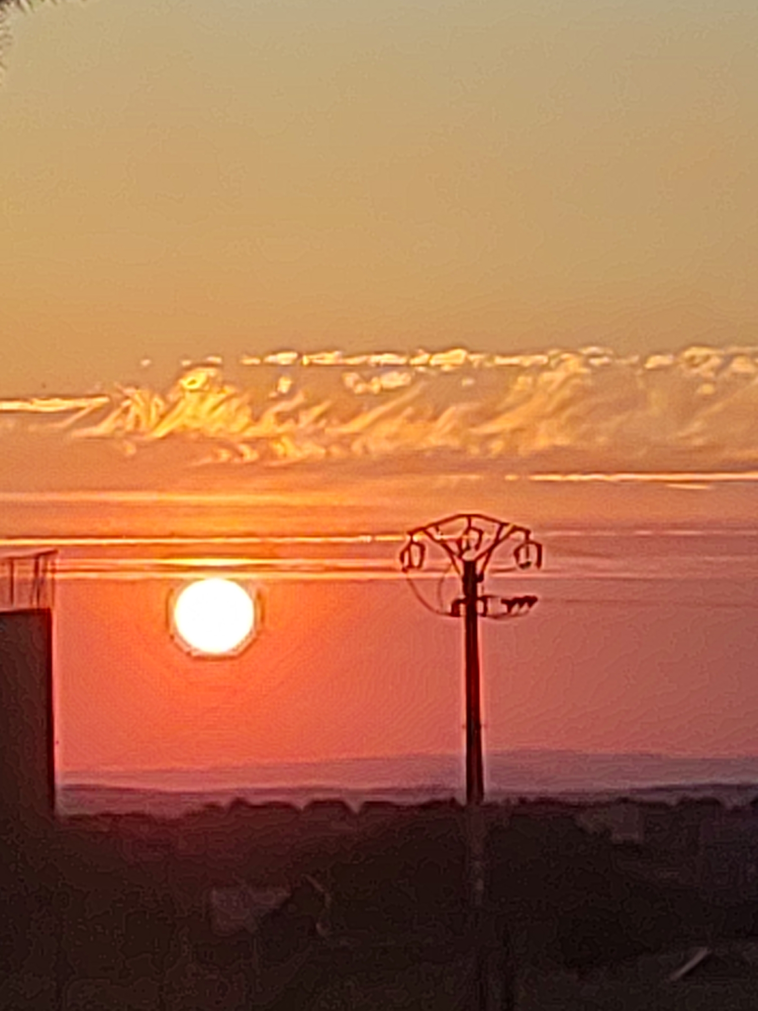 The image depicts a sunset scene in Sobradillo, Spain, characterized by a vibrant orange sky and a bright, octagonal sun partially obscured by clouds. In the foreground, a silhouette of a pylon with multiple arms is visible to the right, and a portion of a building is seen on the left. The landscape below is dark and indistinct, suggesting it is either late evening or night. The overall impression is one of a serene, natural spectacle enhanced by the presence of man-made structures.