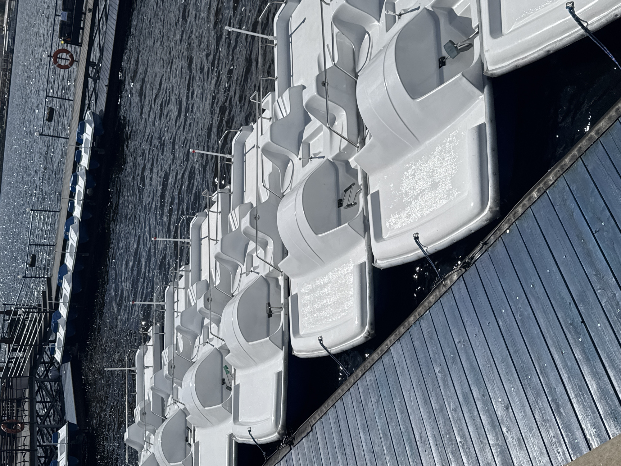 The image shows a row of white pedal boats docked along a blue-gray wooden pier. The boats are lined up parallel to each other, with the pier extending along the right edge of the image. Dark water fills the space to the left of the boats and pier. There is a metal lifeguard donut on a black fence. The image appears to be taken in Izhevsk, Russia.