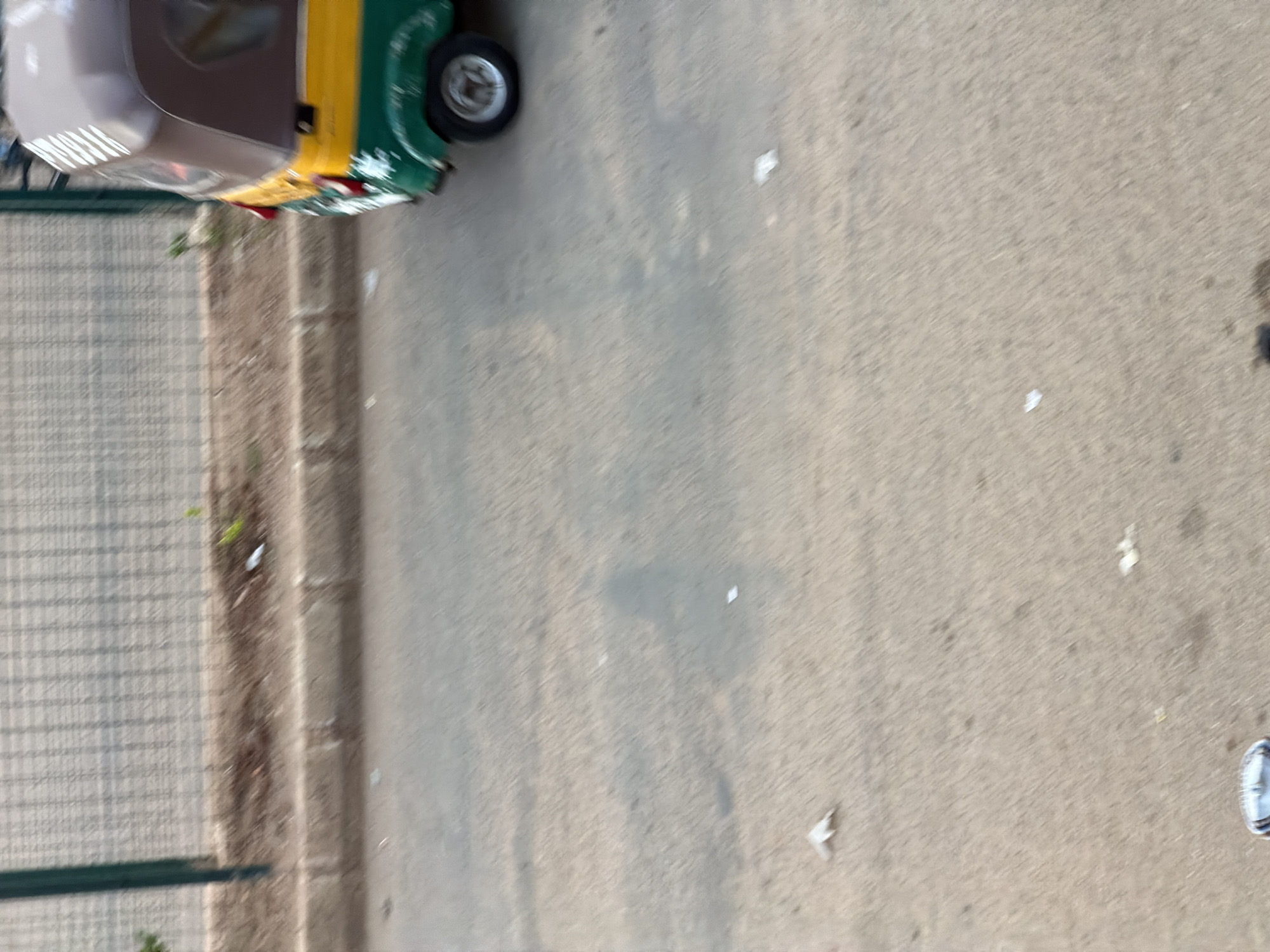 The image depicts a street scene in Bukuru, Nigeria. In the upper right quadrant, a three-wheeled auto-rickshaw, painted green, yellow, and brown, is partially visible. It appears to be in motion, as suggested by a slight blur. The auto-rickshaw is on a paved road. To the left of the road is a dirt verge with sparse vegetation and a wire mesh fence in the background. The road surface is concrete and shows signs of wear, with scattered white debris. The lighting suggests it is daytime. There are no discernible people or specific actions taking place other than the implied movement of the auto-rickshaw. The overall environment is that of an urban or semi-urban street.