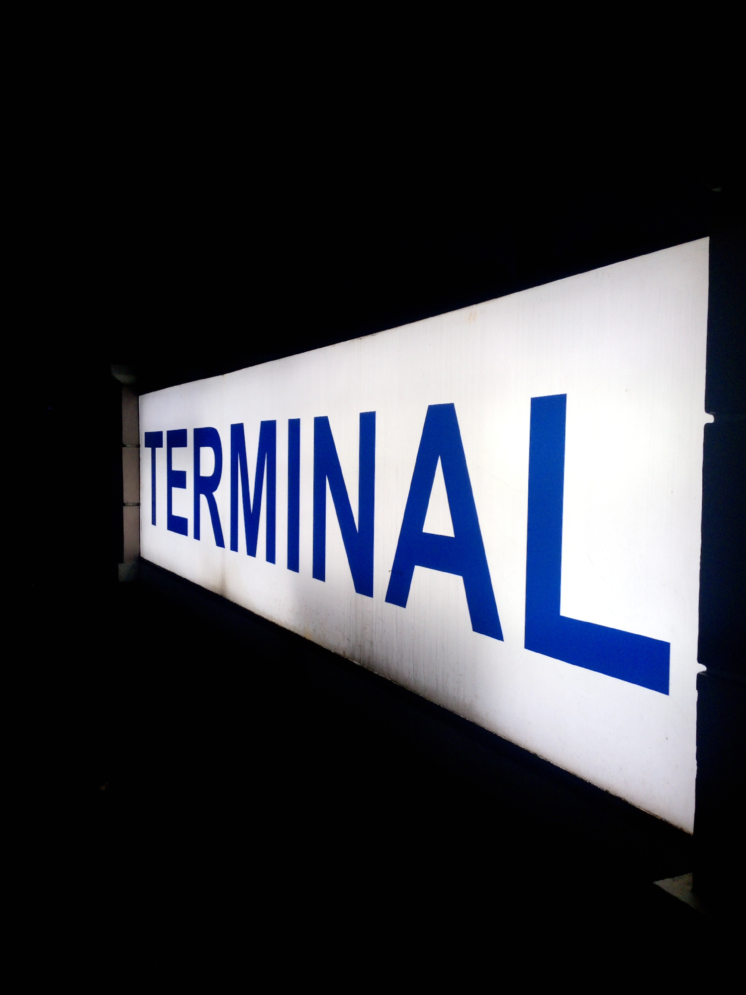 A brightly lit, white sign with blue lettering spelling out "TERMINAL" is angled towards the viewer against a black background. The sign appears to be illuminated from within and is a long, rectangular shape. The lettering is bold and sans-serif. The image is taken from a low angle, with the sign extending into the darkness on both the left and right sides. There are no people or specific environmental details visible beyond the sign and the dark surroundings.