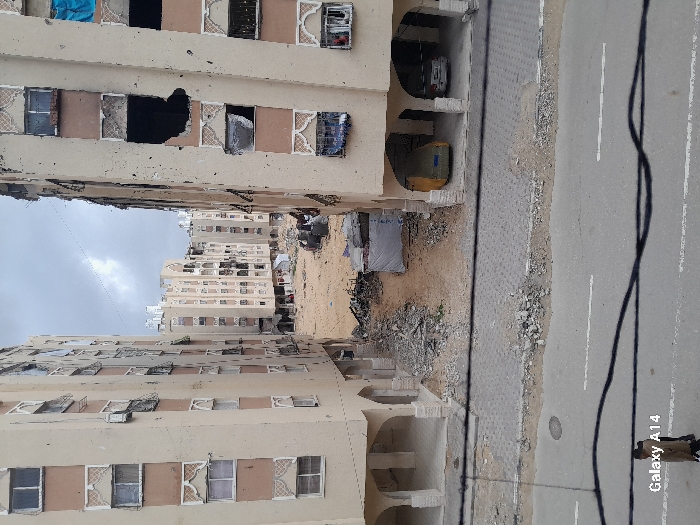 The image presents an elevated view of a severely damaged urban residential area in Khan Yunis, Palestinian Territory, under an overcast sky. Two multi-story, light-colored apartment buildings with brown decorative elements flank a central area of extensive destruction. The building on the right displays significant structural damage, including a large, dark, irregular void on an upper floor and numerous shattered windows, some revealing exposed interiors, one with a blue tarp. A yellow vehicle and a white car are partially visible parked beneath its ground-level arched arcade. The building on the left also shows multiple broken or boarded windows. Between these structures, a large expanse of dirt and sand is covered with considerable rubble, debris, and what appears to be a makeshift shelter under a white tarp, indicating widespread demolition. In the foreground, a paved asphalt road with white double-line markings and a textured concrete sidewalk are visible, with scattered rubble at the edges. Two individuals, appearing to be civilians, walk on the road in the lower-left corner. Utility lines cross the upper foreground.