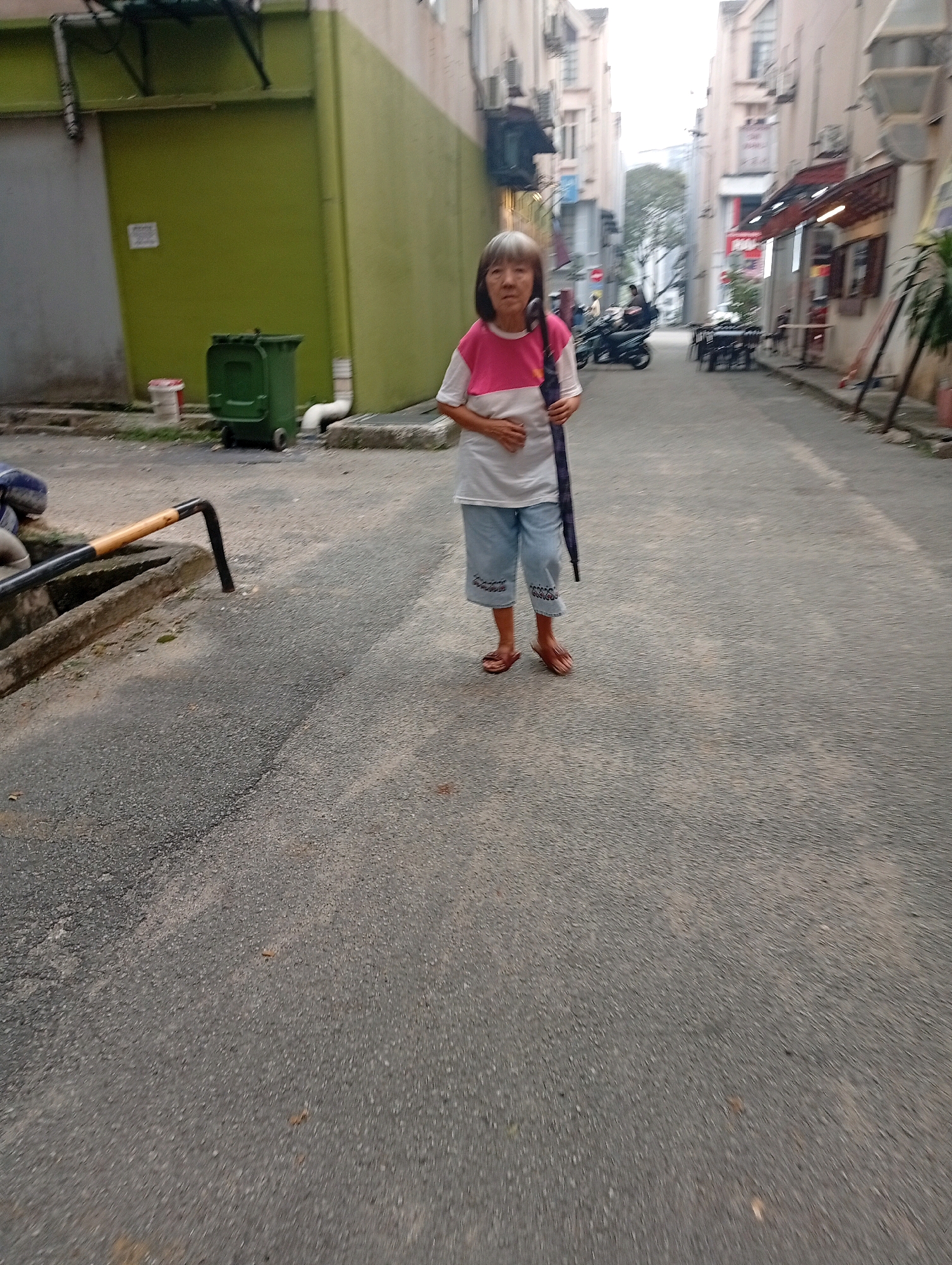 An elderly woman with gray hair stands in a narrow alleyway between buildings. She is wearing a pink and white t-shirt, light blue shorts, and brown sandals. She is holding a dark umbrella vertically in her right hand. The alley has a paved surface and is lined with buildings on both sides. On the left side, there is a green wall, a green trash bin, and a drain with a metal tool. In the background, motorcycles are visible, suggesting a bustling urban environment. The overall lighting indicates it may be daytime, possibly overcast.