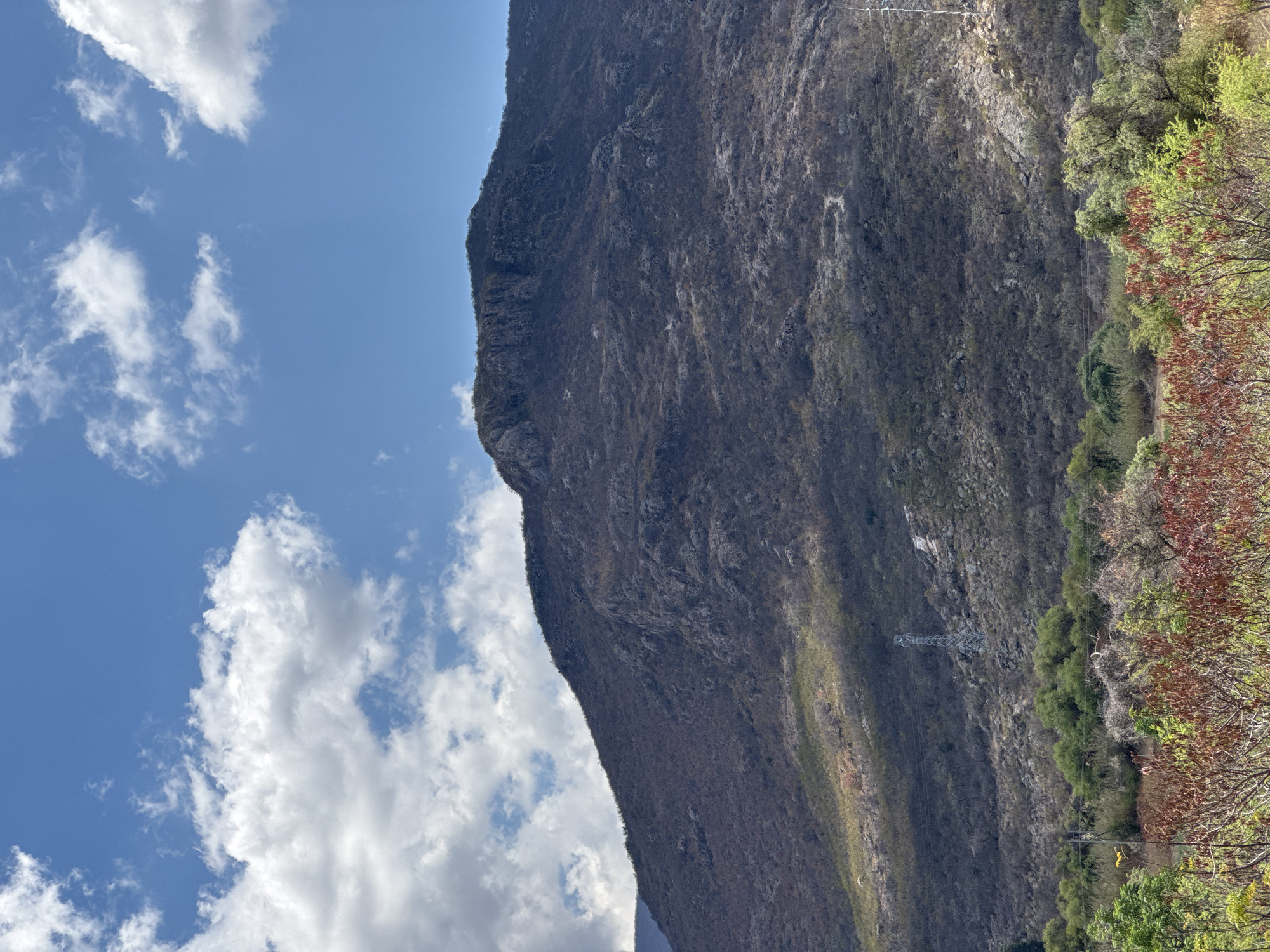 This image depicts a steep mountain face partially covered in sparse vegetation in Tlalixtac de Cabrera, Mexico. The left side of the frame shows a blue sky with white clouds. On the mountain, a power line tower is visible. Vegetation is denser on the right side.