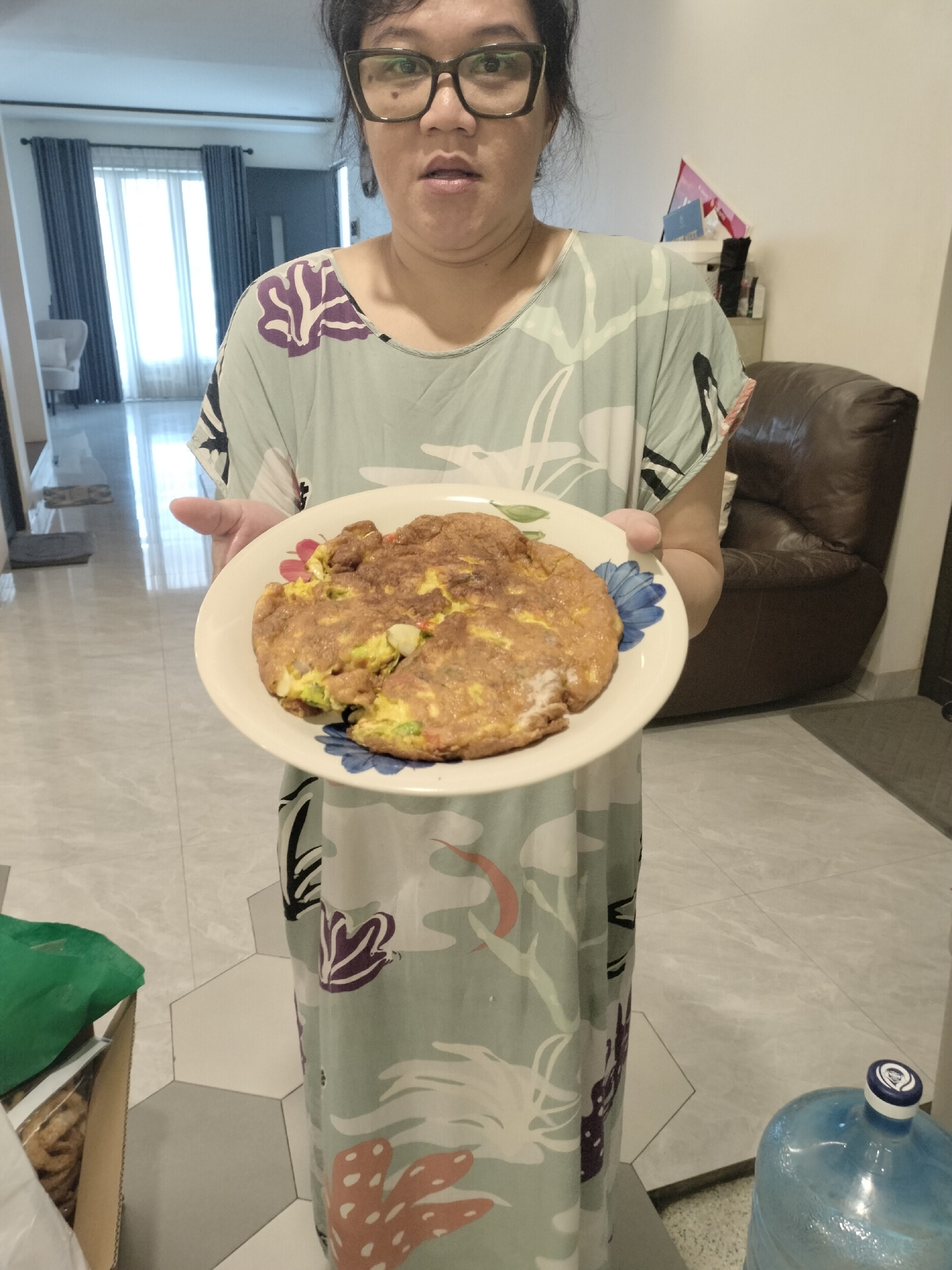 A medium shot captures a woman indoors in what appears to be a residential setting in Bekasi, Indonesia. She is seen from the chest up, holding a white plate with a large, golden-brown omelet or fried egg dish with visible ingredients like vegetables. The plate features blue and red floral patterns.
The woman has dark hair and is wearing black-rimmed glasses, looking directly at the camera with a slightly open mouth and neutral expression. She is dressed in a light-colored, loose-fitting garment with a vibrant floral and abstract pattern in shades of blue, purple, white, and orange.
The background shows a brightly lit room, suggesting daytime. A large window with blue curtains is visible behind her to the left, letting in natural light. To the right, a dark brown leather armchair is partially visible. The floor is covered with light-colored tiles, featuring both square and hexagonal patterns. A large, blue plastic water bottle with a white and blue cap is on the floor to the bottom right. No discernible text is visible on any objects.