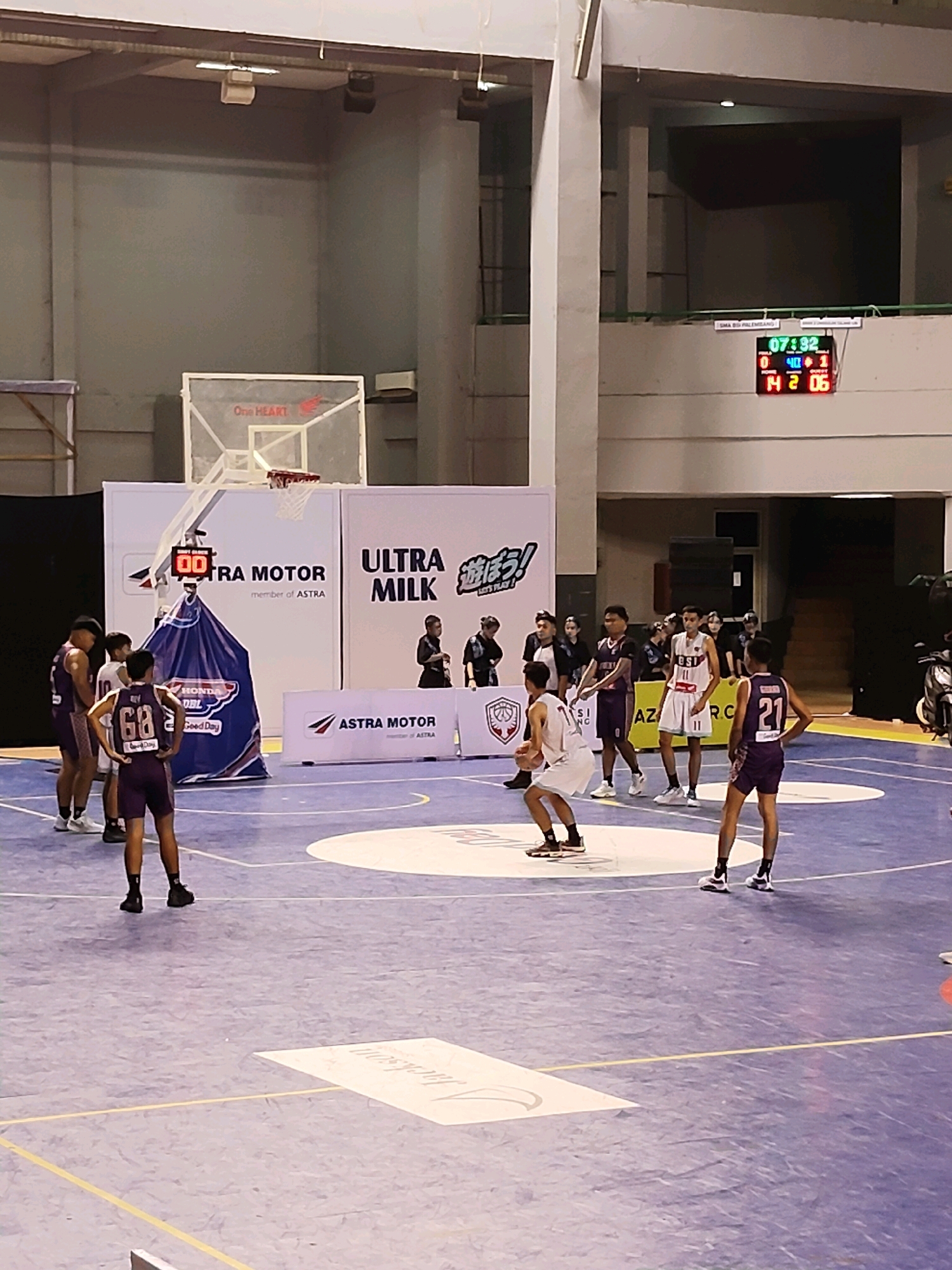 In an indoor basketball arena, a game is underway. The court is a light purple hue with white markings, including a center circle and free-throw lines. Several young men, dressed in basketball uniforms, are on the court. One player in a white uniform with the number 11 is in the foreground, dribbling a basketball. Other players in purple uniforms are scattered around the court, some observing the game, others in defensive positions.
In the background, a large white banner displays "ULTRA MILK" and "ASTRA MOTOR" logos. Behind the players, a scoreboard shows "07:32" as the game time and "0" and "14" as the scores for the respective teams. A basketball hoop and backboard are visible above the banner. Spectators can be seen in the bleachers above the court. The overall environment suggests a competitive sports event.