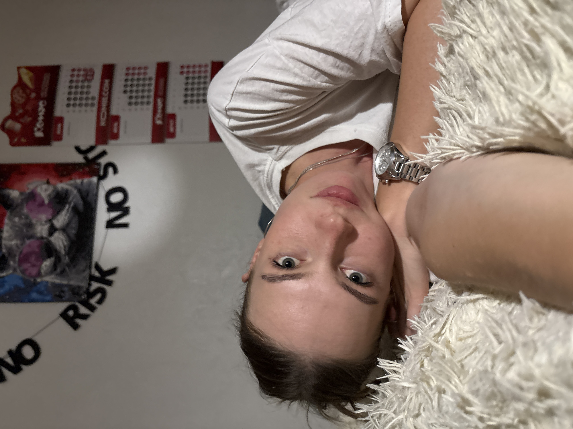 A close-up, eye-level, full-frame shot shows a young woman lying on a fluffy white surface, looking directly at the camera. Her head is tilted to the left, resting on her hand. She has light skin, fair eyebrows, and blue-green eyes. She is wearing a white t-shirt and a silver wristwatch on her left wrist. Behind her, a white wall is visible with a decorative garland of black letters spelling "NO RISK NO STO", partially obscuring a framed picture of a cat wearing pink sunglasses. To the right of the garland, three calendar pages are attached to the wall, each with a red top border and dates displayed in a grid. The overall lighting is dim, suggesting an indoor setting. There are no discernible architectural features or landmarks that would definitively place the scene in Perm, Russia.