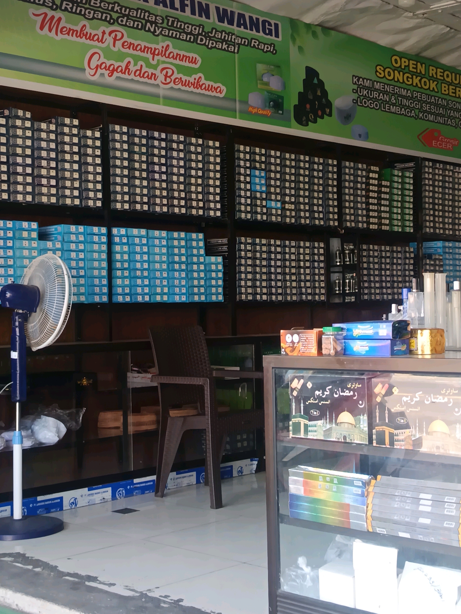 The image shows the interior of a shop, likely in Blega, Indonesia, specializing in songkoks, a type of cap. The shop is well-stocked, with numerous shelves filled with boxed songkoks in various sizes and designs. A large green banner at the top advertises the shop, "ALFIN WANGI," and highlights the quality of their products with phrases like "Berkualitas Tinggi, Jahitan Rapi, Ringan, dan Nyaman Dipakai" (High Quality, Neat Stitching, Light, and Comfortable to Wear). The banner also offers customization options, such as "UKURAN & TINGGI SESUAI YANG DIINGINKAN" (Size & Height As Desired) and "LOGO LEMBAGA, KOMUNITAS, ORGANISASI" (Logo for Institutions, Communities, Organizations).
In the foreground, a blue pedestal fan is visible, suggesting a warm climate. A glass display case contains boxes with Arabic script, possibly related to Ramadan or religious items. The shelves are neatly organized, and the overall impression is of a clean and professional retail environment. The lighting appears to be artificial, likely from ceiling lights, illuminating the products and the shop's interior. No people are clearly visible in the image, but the presence of merchandise indicates active commerce.