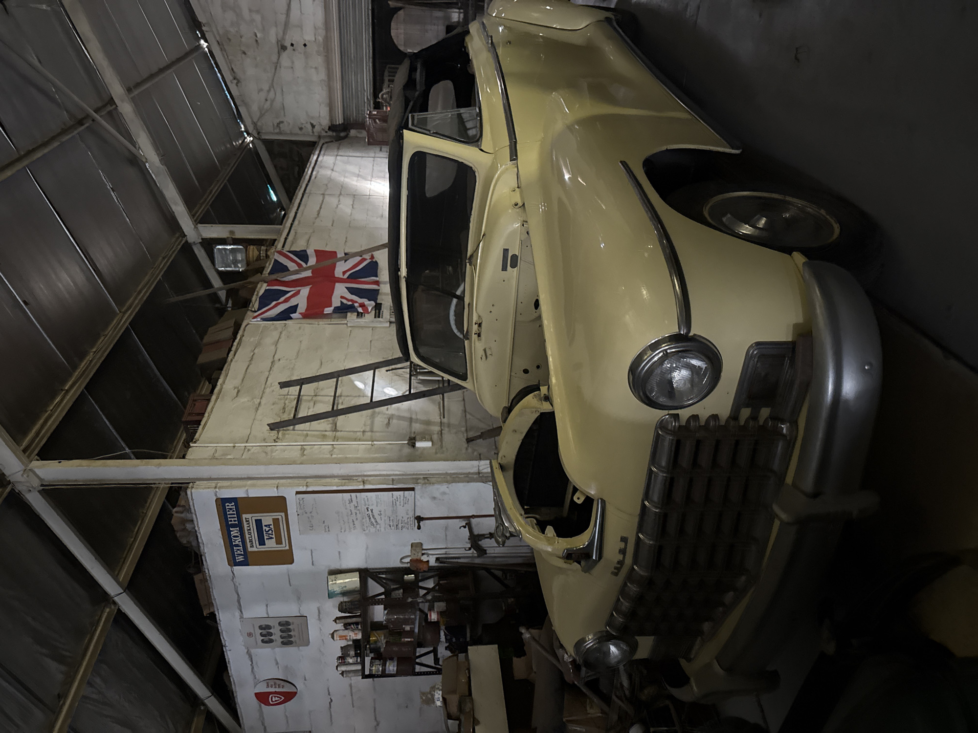 The image depicts a light yellow vintage car, possibly in a state of restoration or disrepair, inside a garage or workshop. The car's door is open, revealing part of the interior. Behind the car, a British Union Jack flag is hanging on the wall. A ladder leans against the wall as well. Other items are visible, including shelves with various containers and cans, and signage on the wall. The environment appears to be a cluttered, industrial-style space. The location could be in Kraaifontein, South Africa, as indicated by the "Welkom Hier" (Welcome Here) signage in Afrikaans and the reference to a Barclayskaart Visa card. There is no indication of people visible in the photo.