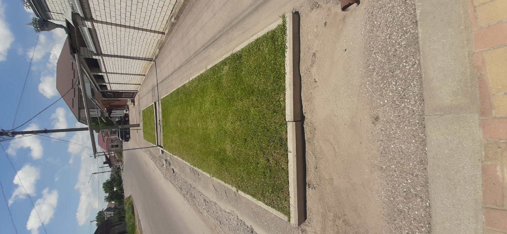 The image depicts a street scene in Yessentukskaya, Russia, on a sunny day with a partly cloudy sky. In the foreground, a well-maintained rectangular patch of bright green grass is bordered by concrete curbs. Behind this grass patch, another smaller, similar grass area is visible. A paved walkway runs alongside the grass and extends into the background. Adjacent to the walkway is a white brick wall topped with decorative elements. To the left of the walkway and grass areas, a gray asphalt road curves into the distance. Along the road, several buildings and houses are visible, with trees and vegetation in the background. A utility pole with numerous wires stands prominently near the center of the image, extending upwards towards the sky. A dark-colored car is parked along the roadside further down the street, near a building with a pink facade. The overall environment suggests a suburban or residential area within the city.
