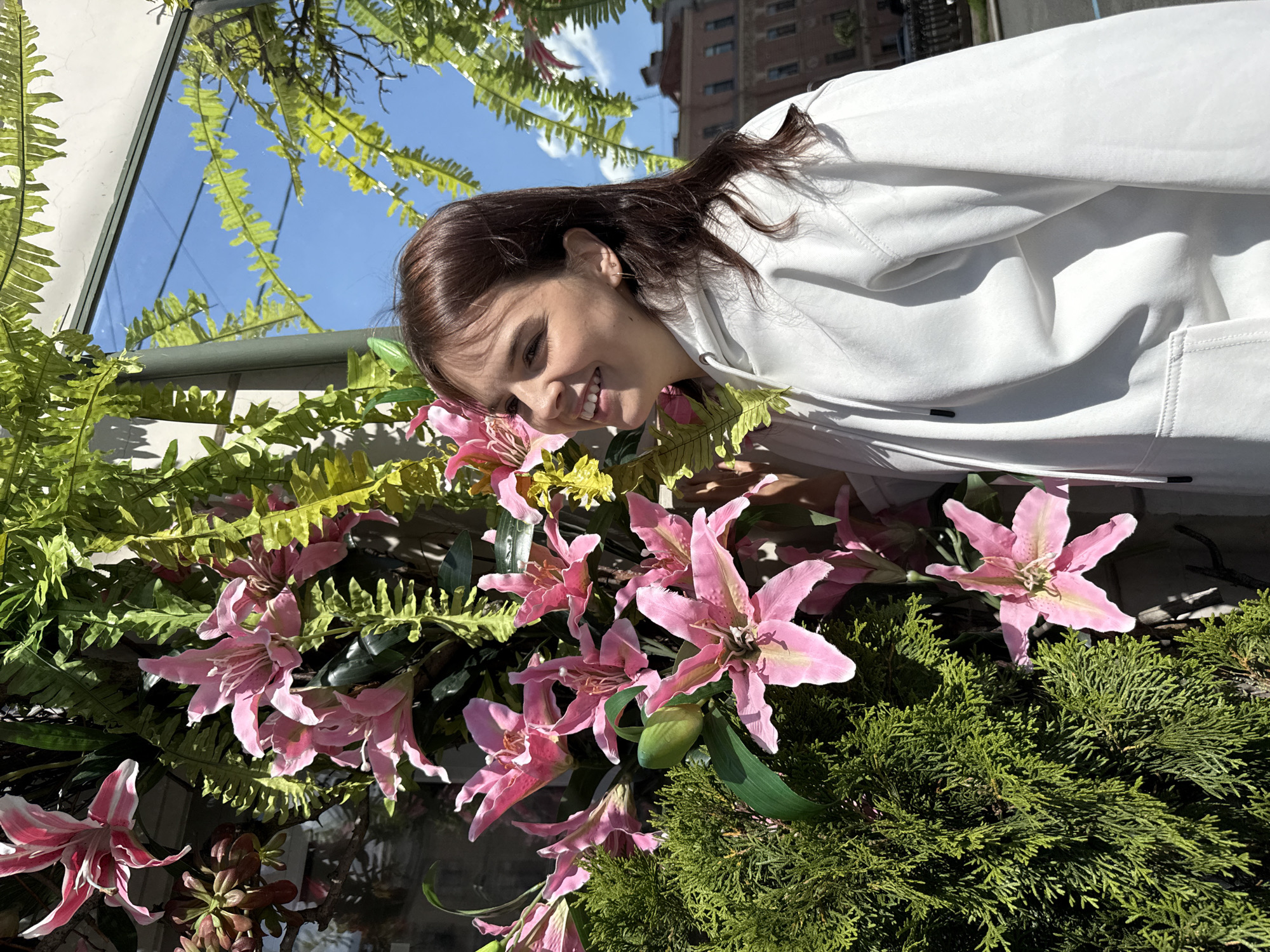 A young woman with brown hair stands in front of a display of artificial flowers and greenery, her head turned to the right in a smiling profile. She is wearing a white hooded sweatshirt. The display includes large pink lilies and fern fronds. Behind her, a building with architectural details is visible, suggesting an urban setting in Saint Petersburg, Russia. The sky is blue with some clouds. The overall impression is a bright, sunny day.