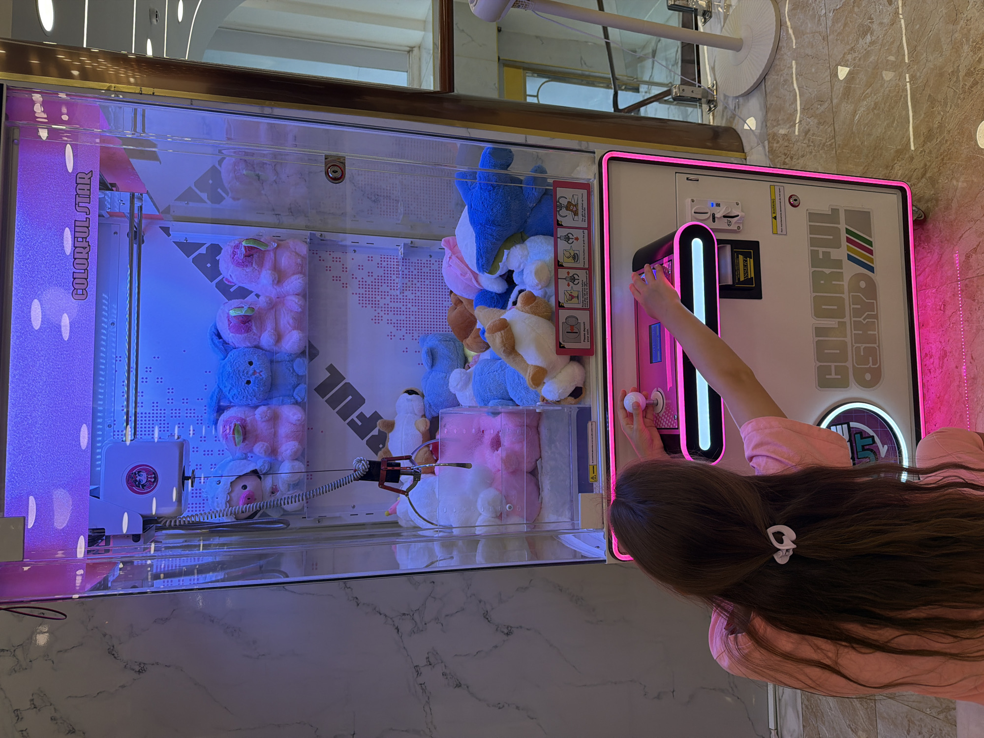 A young girl with long brown hair tied back with a white scrunchie is playing a claw machine in what appears to be a mall or entertainment center. The claw machine is illuminated with pink and blue lights, and the words "COLORFUL STAR" are visible at the top. Inside the machine, there are various plush toys, mostly in pastel colors. The girl is holding a joystick, her hands positioned over the control panel. The environment appears to be indoors with marble flooring and a wall to the left that resembles marble. The location is identified as Nha Trang Ward, Vietnam.