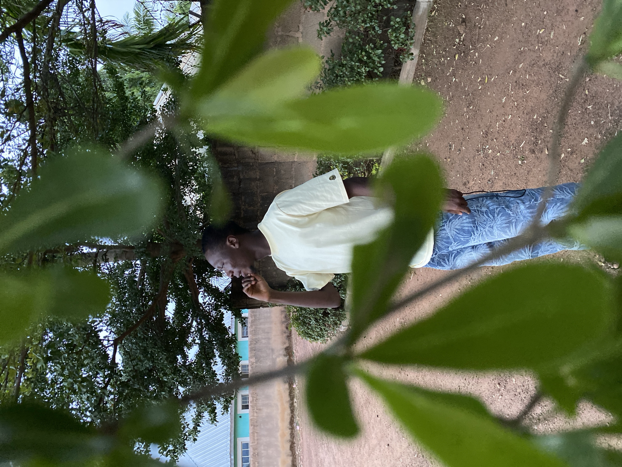 A medium, slightly overexposed, side-view shot shows a man standing outdoors, possibly in Bukuru, Nigeria, based on environmental factors and inference. The man is dark-skinned with short hair, wearing a light yellow, short-sleeved shirt and light blue jeans with a leafy or floral pattern. He appears to be looking down and possibly bringing something to his mouth with his right hand. The foreground is heavily obscured by out-of-focus green foliage. In the background, behind the man, a building with multiple windows and a light-colored (possibly blue-green) exterior can be seen. Beyond the building, there are trees and a tan-colored wall. To the right of the man, there is a dirt or gravel ground surface with scattered leaves. The image is oriented with the right side of the photo facing up and the left side facing down.