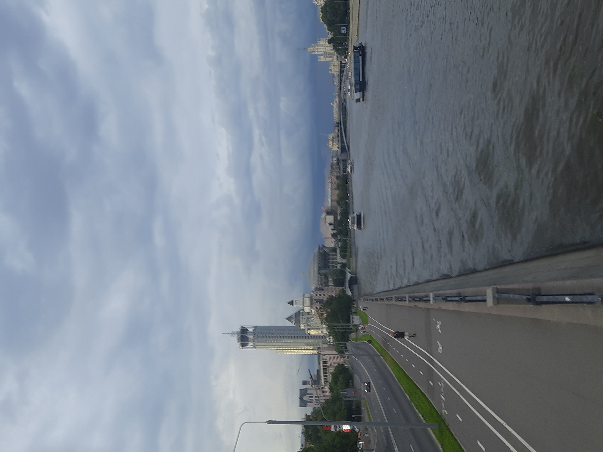 The image depicts a cloudy day scene of a city riverfront. In the foreground, a paved walkway runs alongside a road with a bike lane. A lone male figure is walking on the walkway. The river is to the right of the walkway, with several boats visible on its surface. In the background, a cityscape with various buildings, including a tall skyscraper, is visible. A bridge spans the river in the distance, and a large, distinctive white building with a spire is seen on the far right. The overall impression is that of an urban environment on a slightly overcast day.