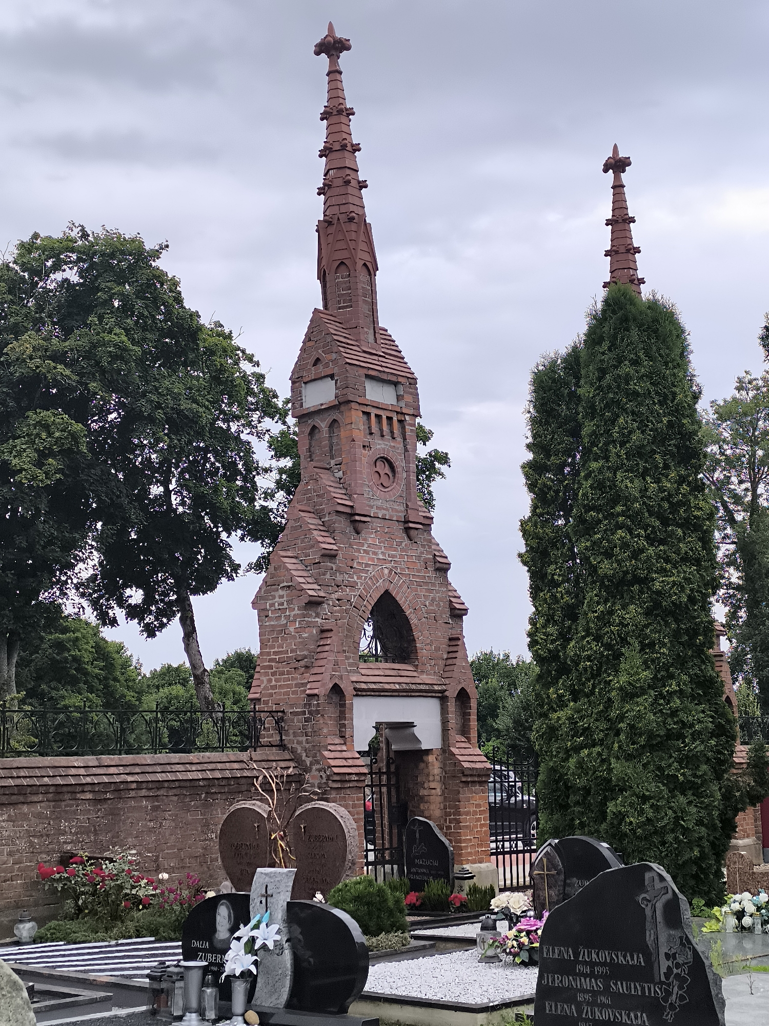 The image depicts a cemetery in Kretinga, Lithuania, featuring a prominent brick mausoleum with two tall spires. The mausoleum is the central focus, constructed from red brick with Gothic architectural elements, including arched openings and decorative finials on the spires. Surrounding the mausoleum are gravestones and burial plots, some with flowers and religious symbols. Evergreen trees and deciduous trees are visible in the background, creating a natural backdrop. The sky is overcast, suggesting a cloudy day. The scene is peaceful and solemn, typical of a cemetery environment.