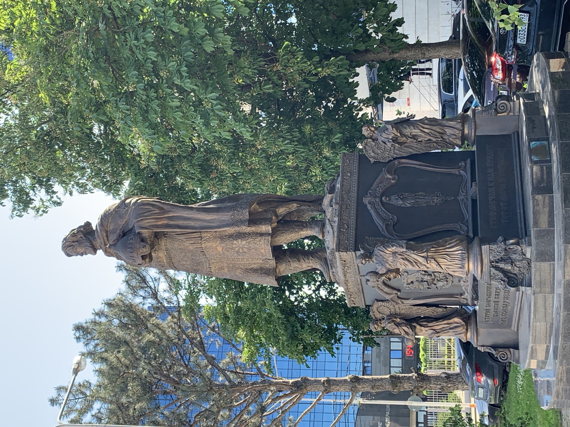 The image shows a statue of a regal figure standing on an ornate plinth, with several smaller figures in relief around the base. The statue is made of bronze or a similar metal. Trees surround the statue, and a modern glass-fronted building is visible in the background. Cars are parked nearby. Text on the monument includes Russian words, including "ФЕДЕРАЦИИ," "ВЕЛИКИЙ КНЯЗЬ," and "АЛЕКСАНДР." A digital display showing "154" is also visible. The license plate of one of the cars, a black Toyota, partially reads "0033CO 93".
