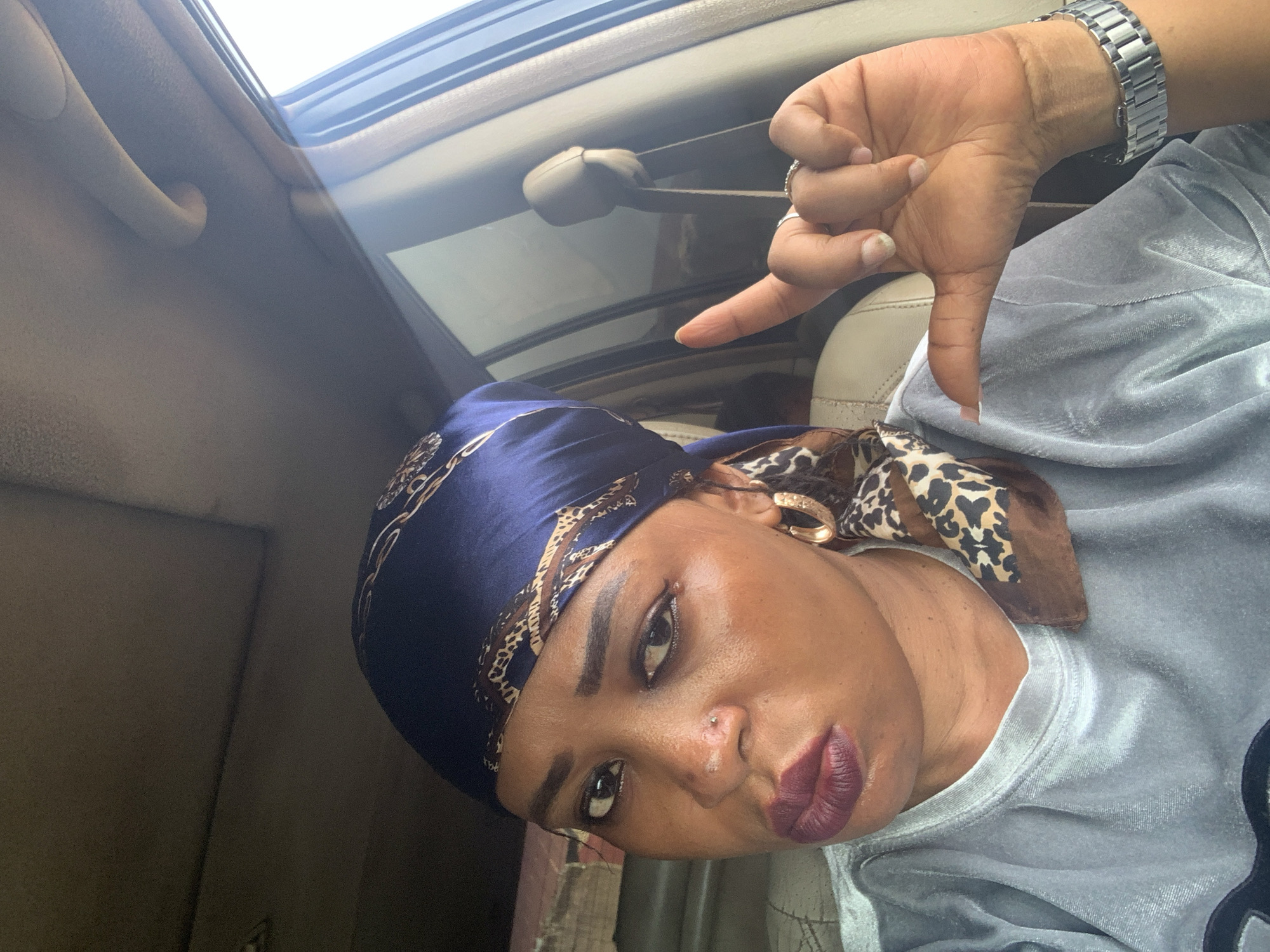 A woman inside a car with a sunroof points downwards with her thumb. The woman is wearing a dark blue headscarf patterned with gold designs and a leopard-print piece of fabric tied at the side. She has gold hoop earrings, dark lipstick, and is wearing a gray shirt. The car's interior is beige. The location is unknown.
