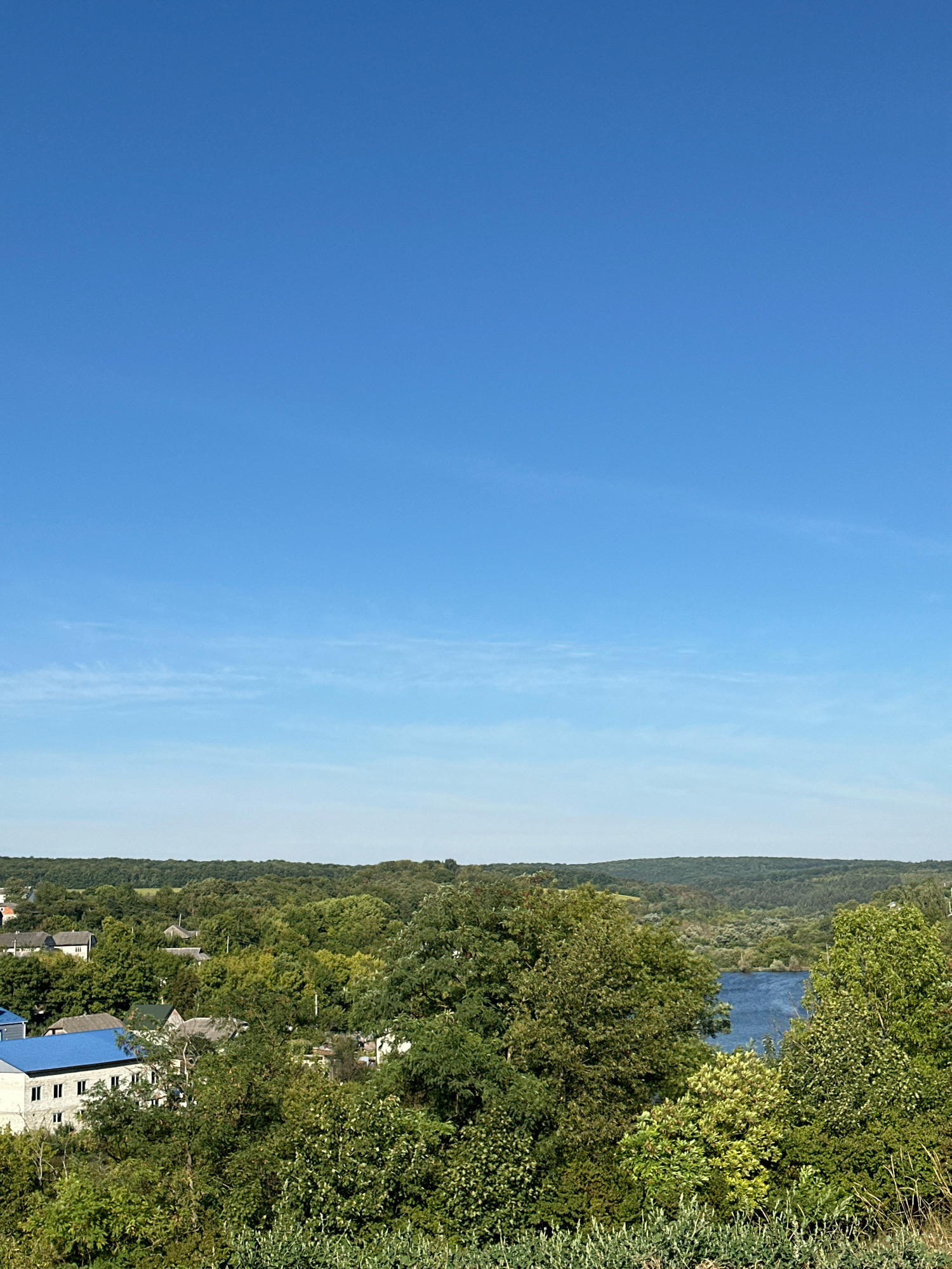 The image depicts a rural landscape under a clear blue sky with a few wispy clouds. In the foreground, a dense array of lush green trees and foliage dominates the lower portion of the frame. Among the trees, several buildings are visible. A prominent white building with a bright blue roof is situated on the left, featuring multiple windows. Further into the scene, scattered houses with gray roofs are nestled amongst the trees, suggesting a small village or settlement. In the middle ground, a body of blue water, likely a river or lake, is visible, bordered by undulating green hills and dense forests that recede into the distance. The scene appears to be a serene, natural setting within or near the city of Makiv, Ukraine.